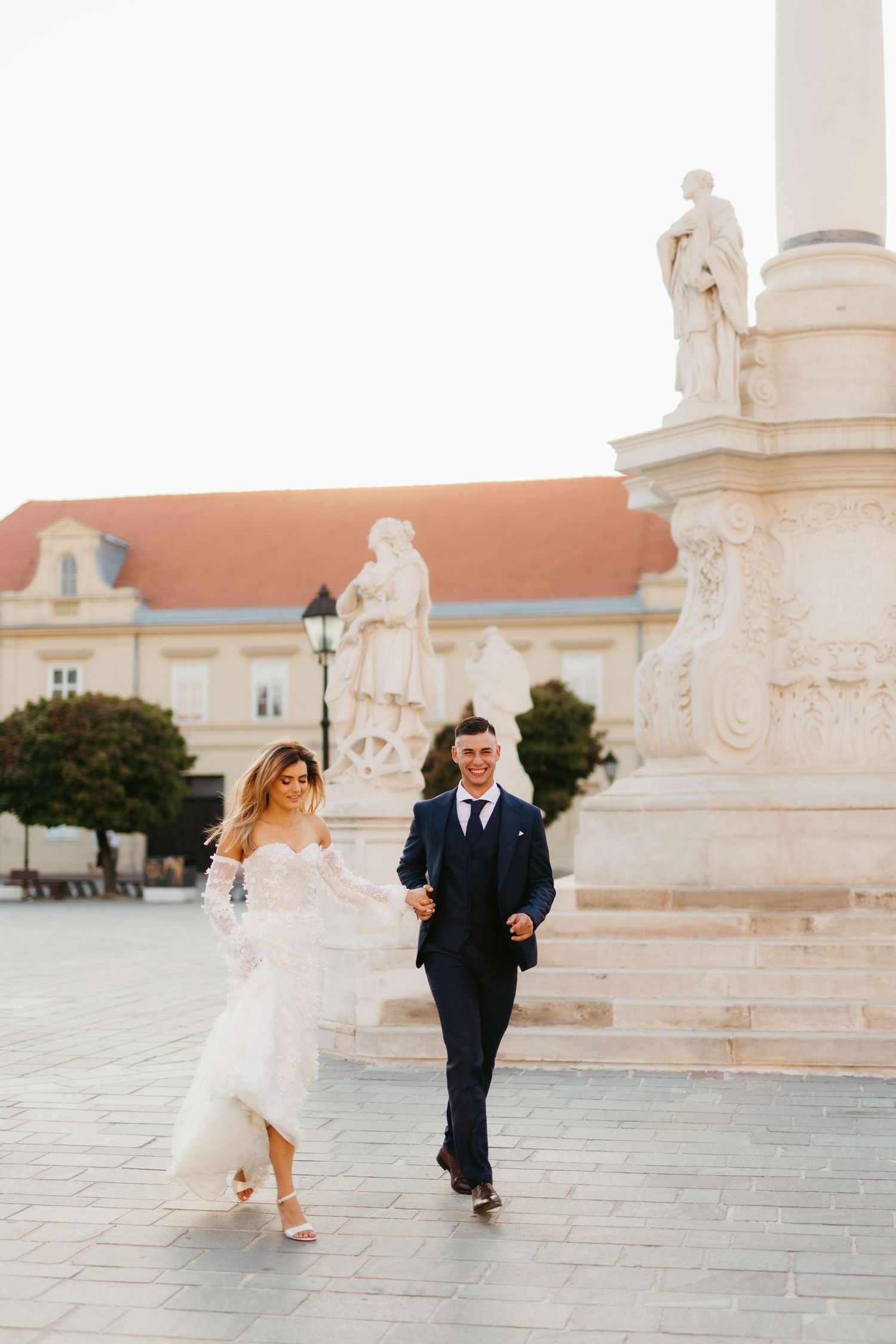A couple in wedding attire runs playfully across a stone courtyard.