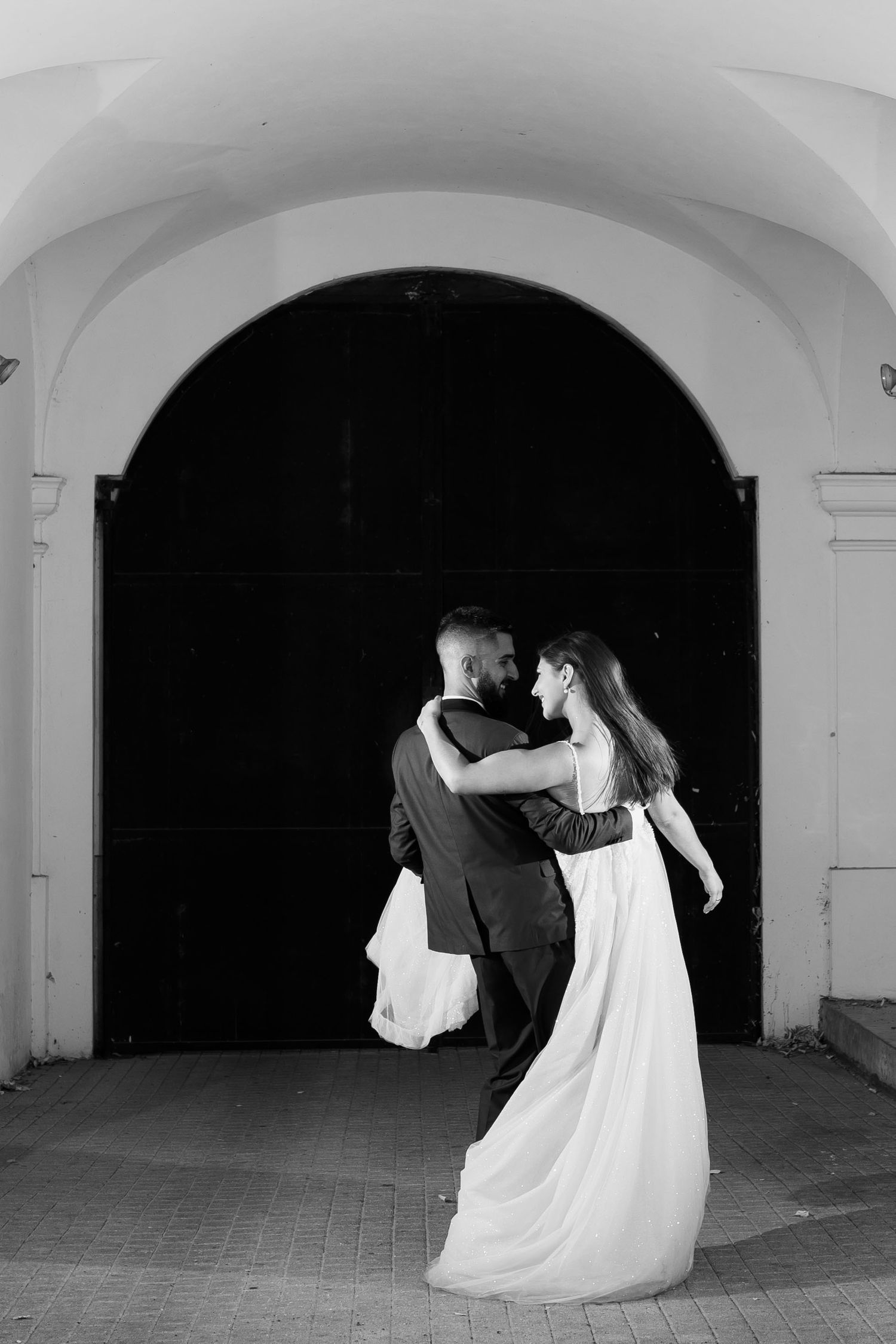 A couple in wedding attire share a romantic dance moment under an arched doorway in black and white photography.