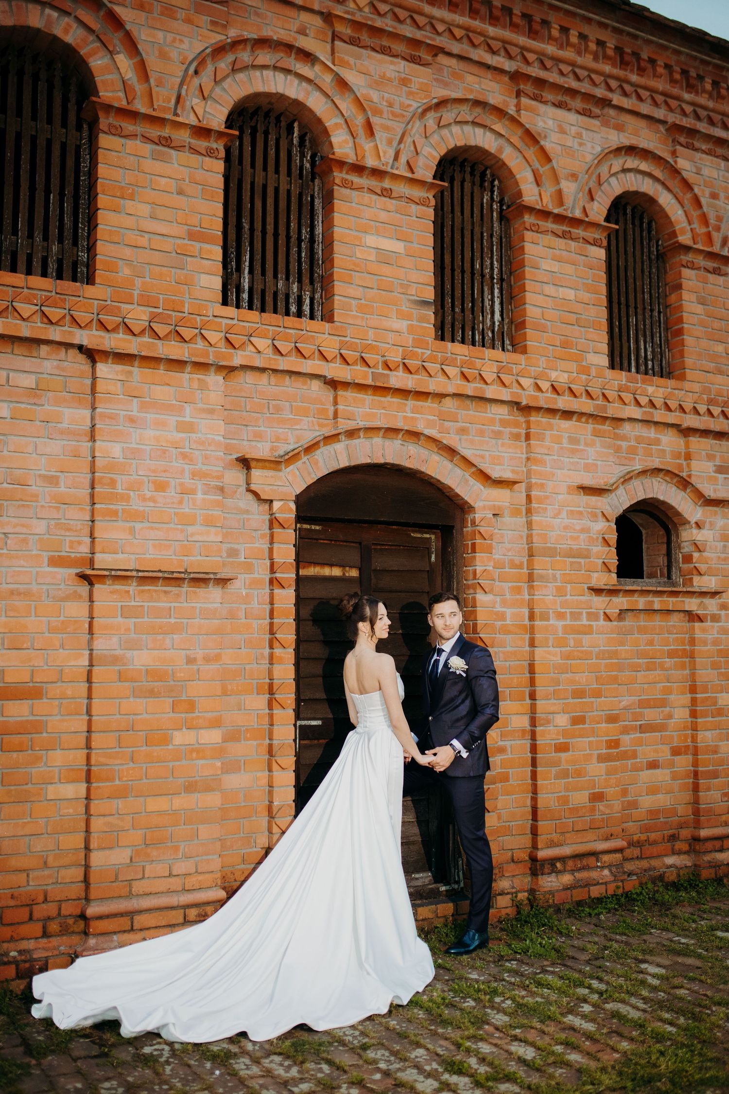 Elegant wedding photo in front of rustic brick building with arched windows and ornate architectural details.