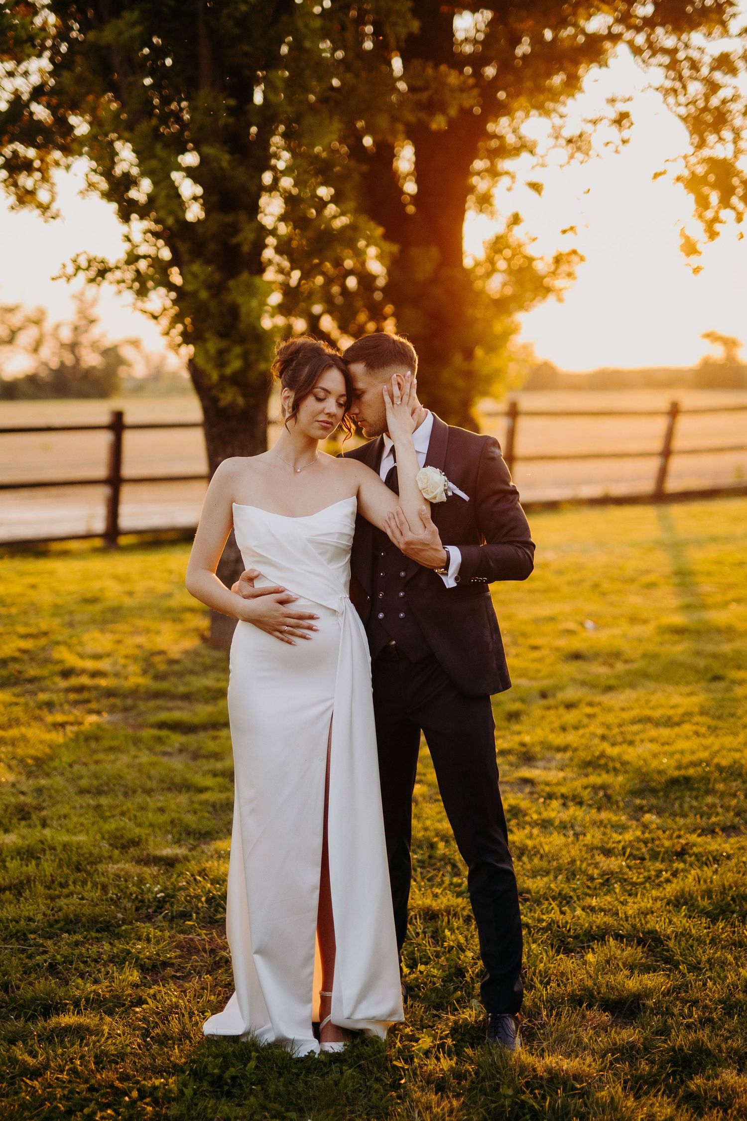 A romantic sunset wedding photo shows a couple in formal attire embracing on a grassy field with wooden fences.