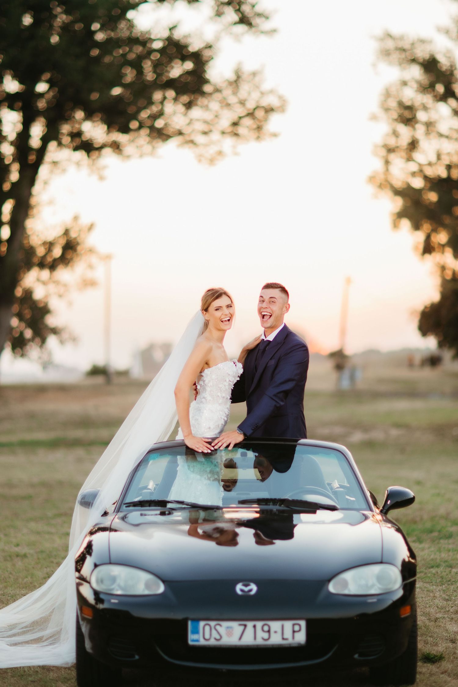 Joyful wedding couple poses on their black Mazda Miata during golden hour photography session.