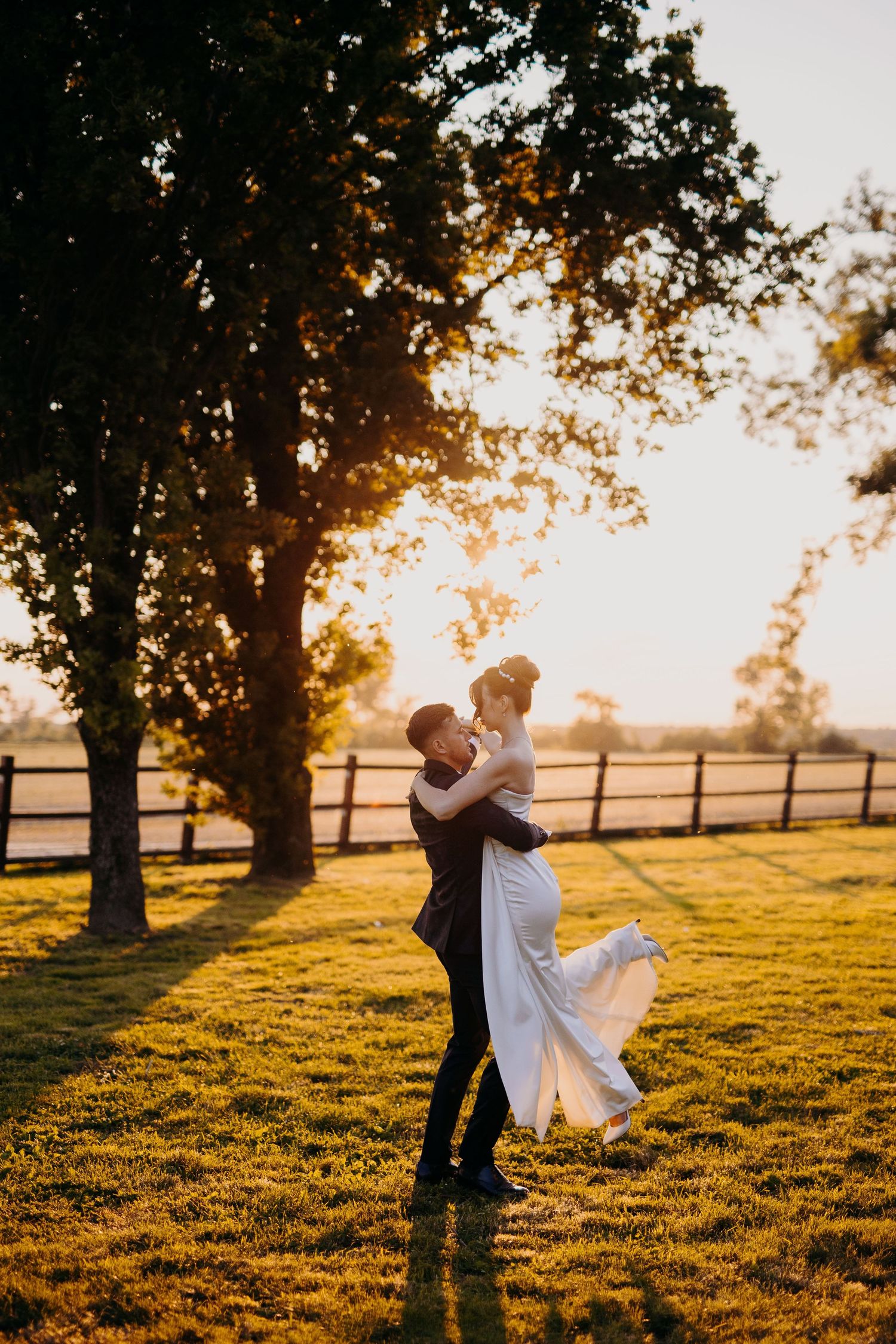 Newlyweds embrace in sunset light on grassy field with wooden fence and trees in background.