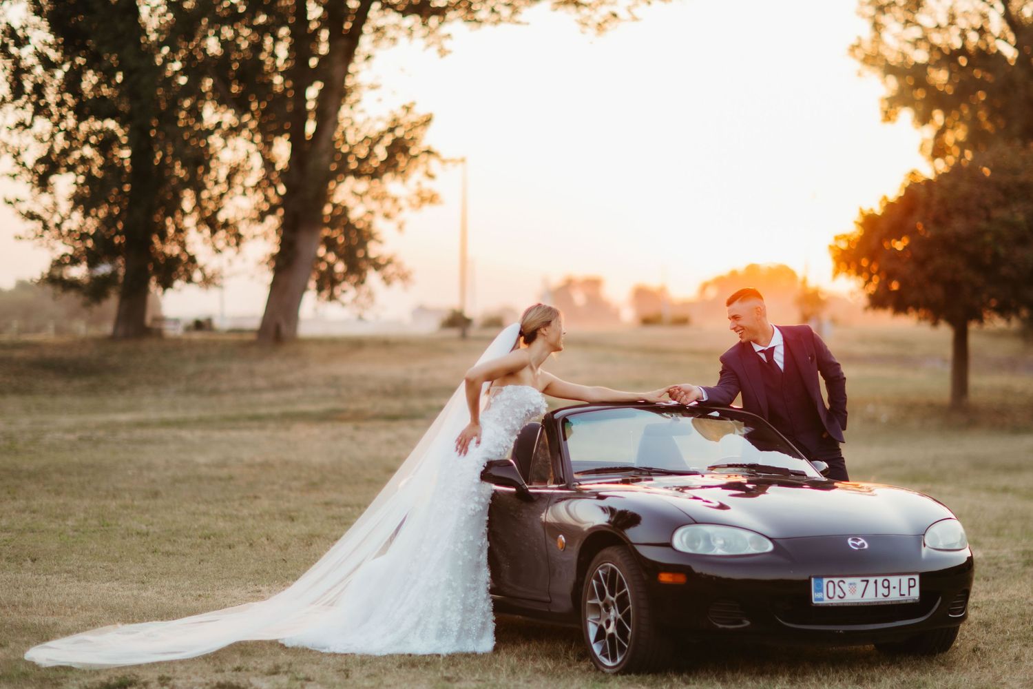 Newlyweds pose with their black Mazda Miata convertible during golden hour sunset in a grassy field.