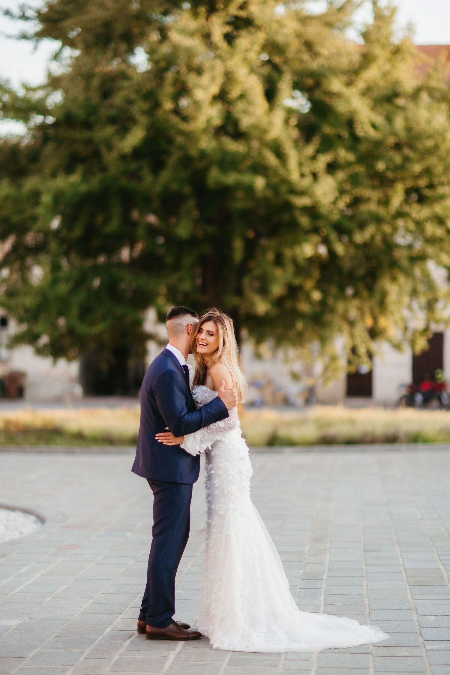 Couple embracing in elegant wedding attire on a historic city square.