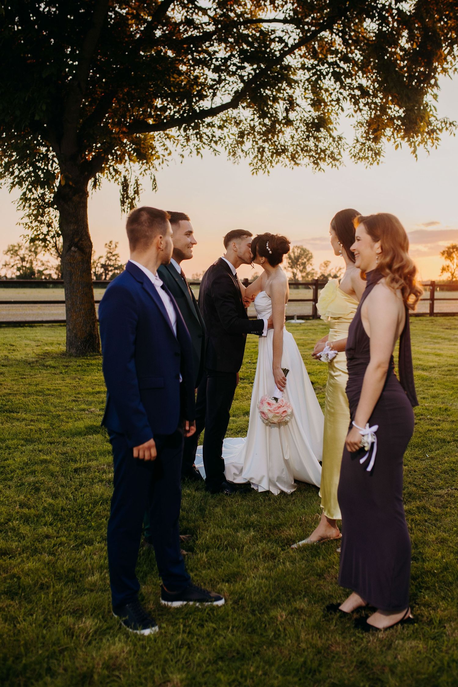 Wedding party gathers on grassy lawn at sunset during outdoor celebration ceremony.