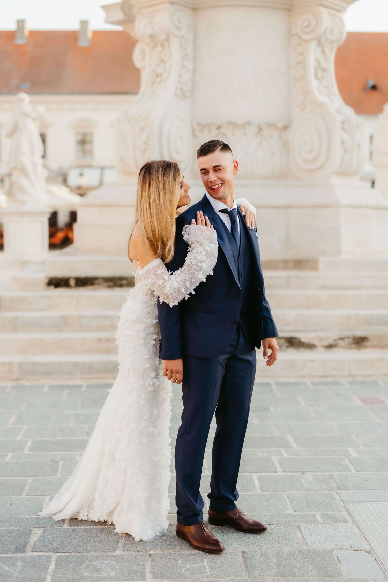 A romantic wedding couple embraces by an ornate white stone column in European architecture setting.