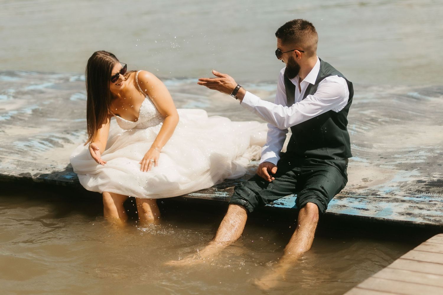 A romantic beach photoshoot of a couple in wedding attire sitting at the edge of water with waves splashing over them.