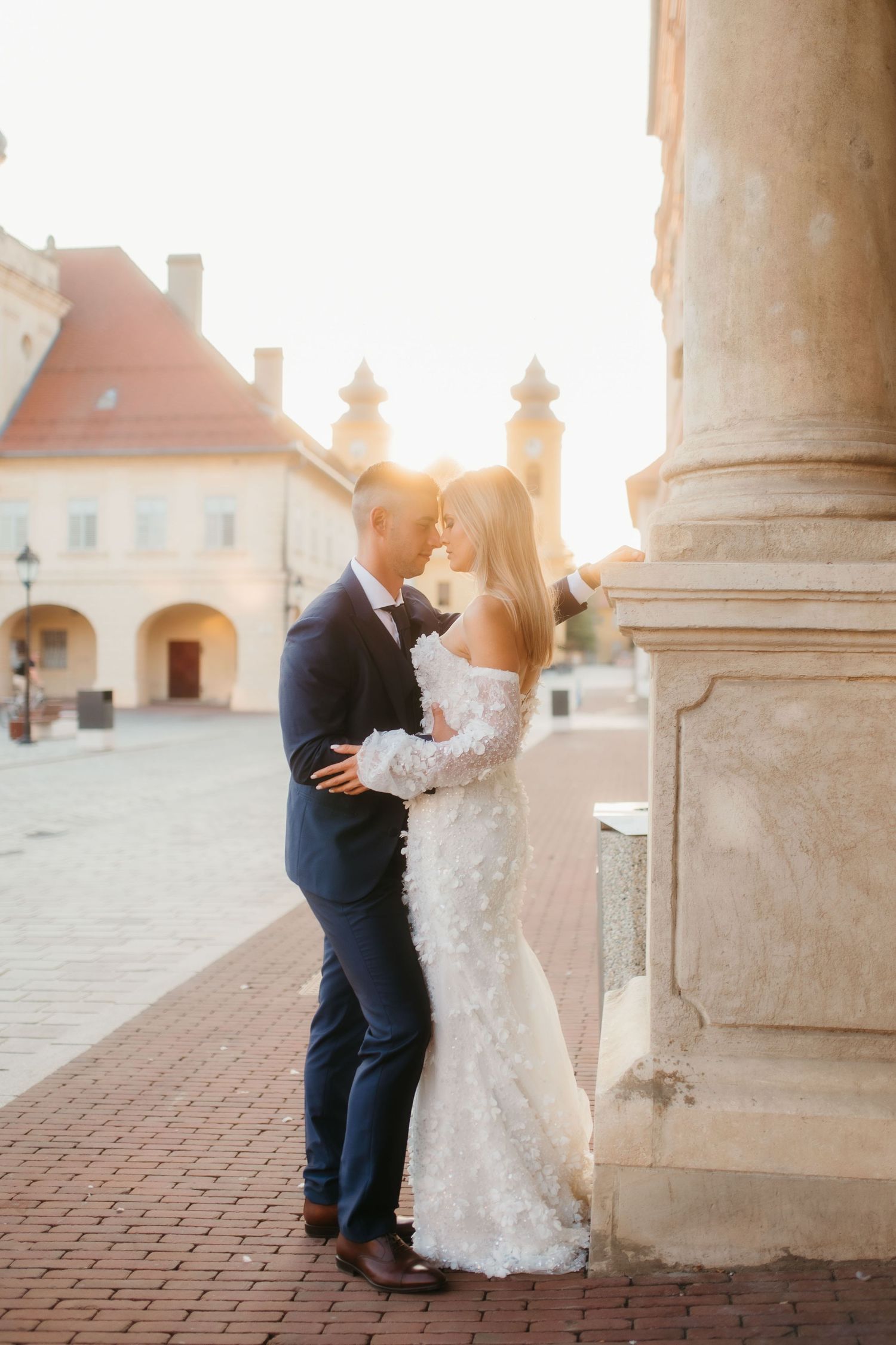Newlyweds embrace by a classic European building with sunlight streaming through stone columns.