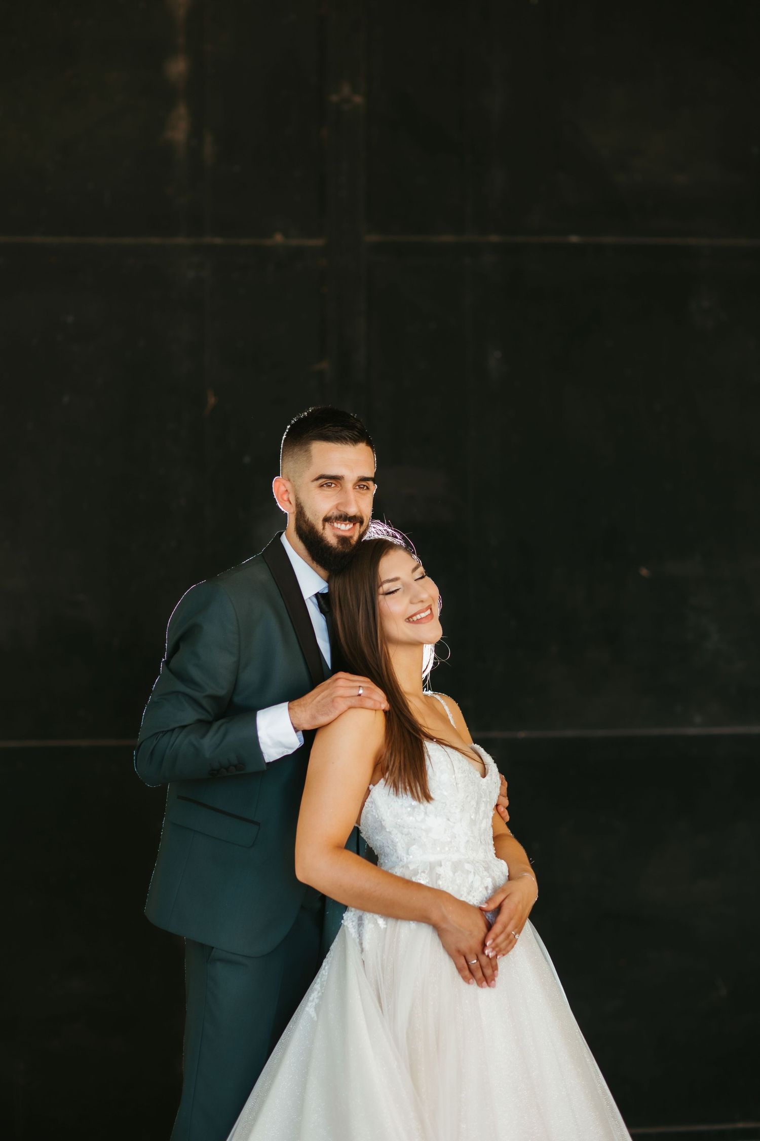A couple in wedding attire embrace against a dark wall, with the bride in white and groom in a teal suit.