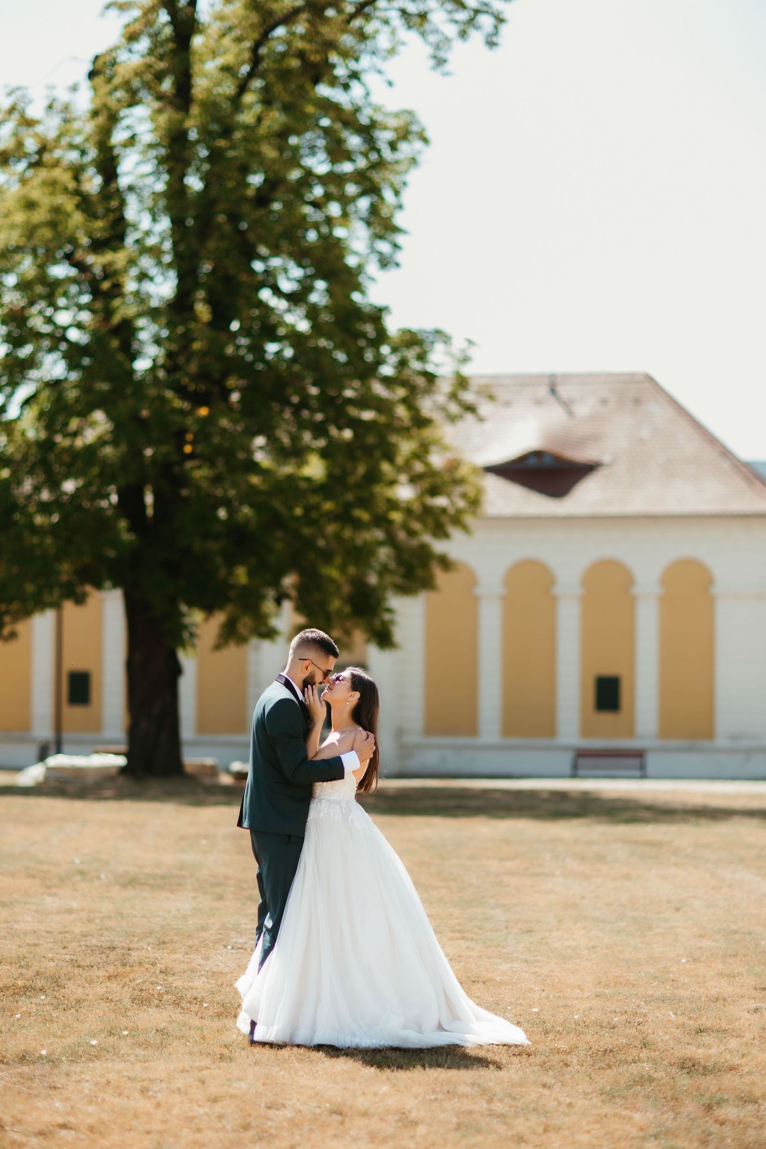 A couple shares an intimate wedding moment on a outdoor lawn near a yellow building with arched windows and large tree.