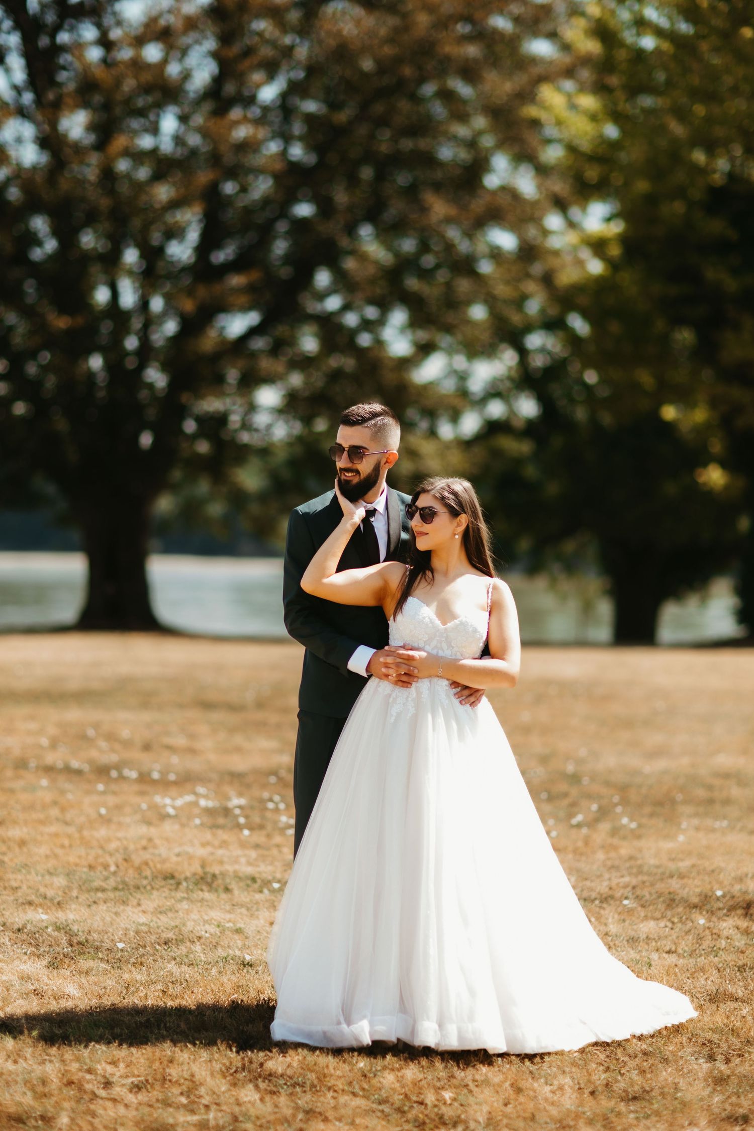 A bride in a flowing white gown and groom in a dark suit pose together on a sunlit lawn with autumn trees in background.