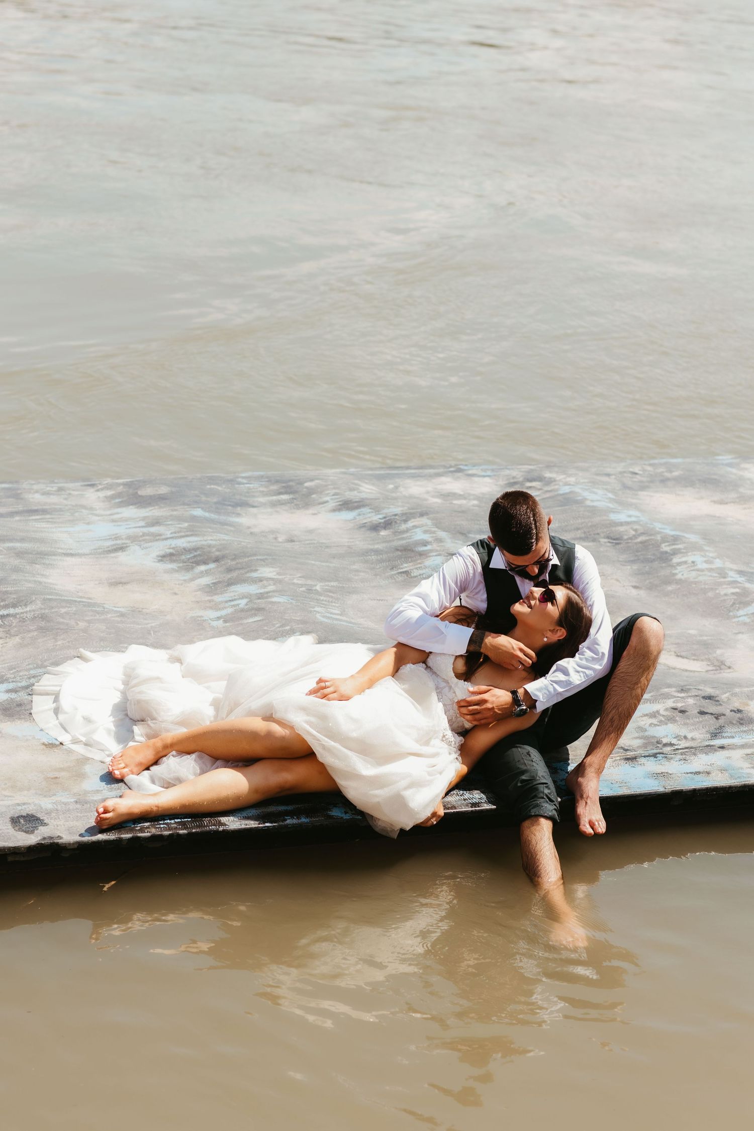 A romantic couple in formal attire embrace while lying on a jetty at the water's edge.