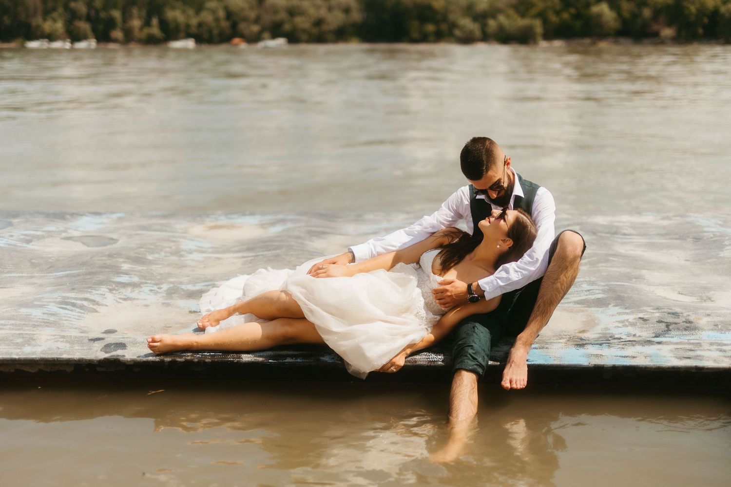 A couple in wedding attire sits together on the edge of a scenic shoreline during their romantic photo session.