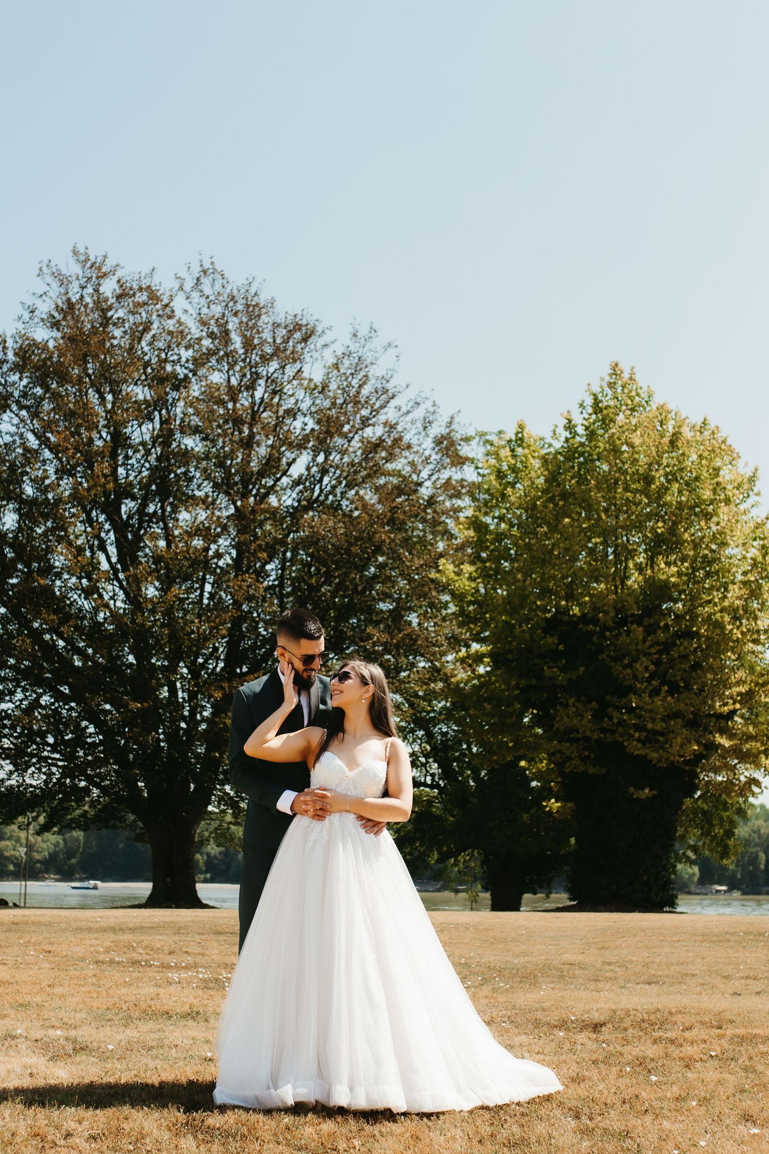 Two people in white wedding dresses standing together in a sunlit field with large trees in the background during golden hour.