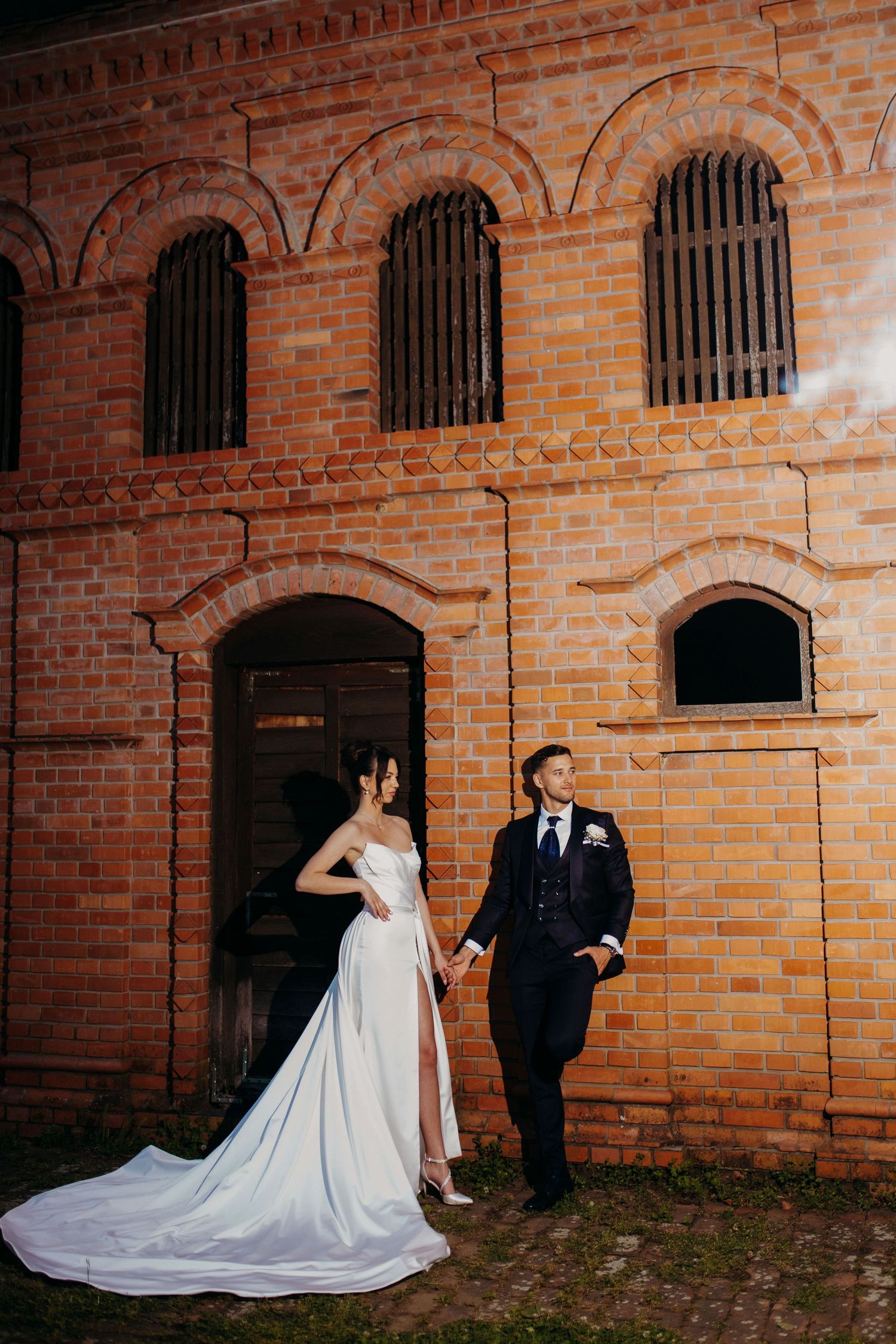 A couple poses in wedding attire against a rustic brick building with arched windows and iron bars.