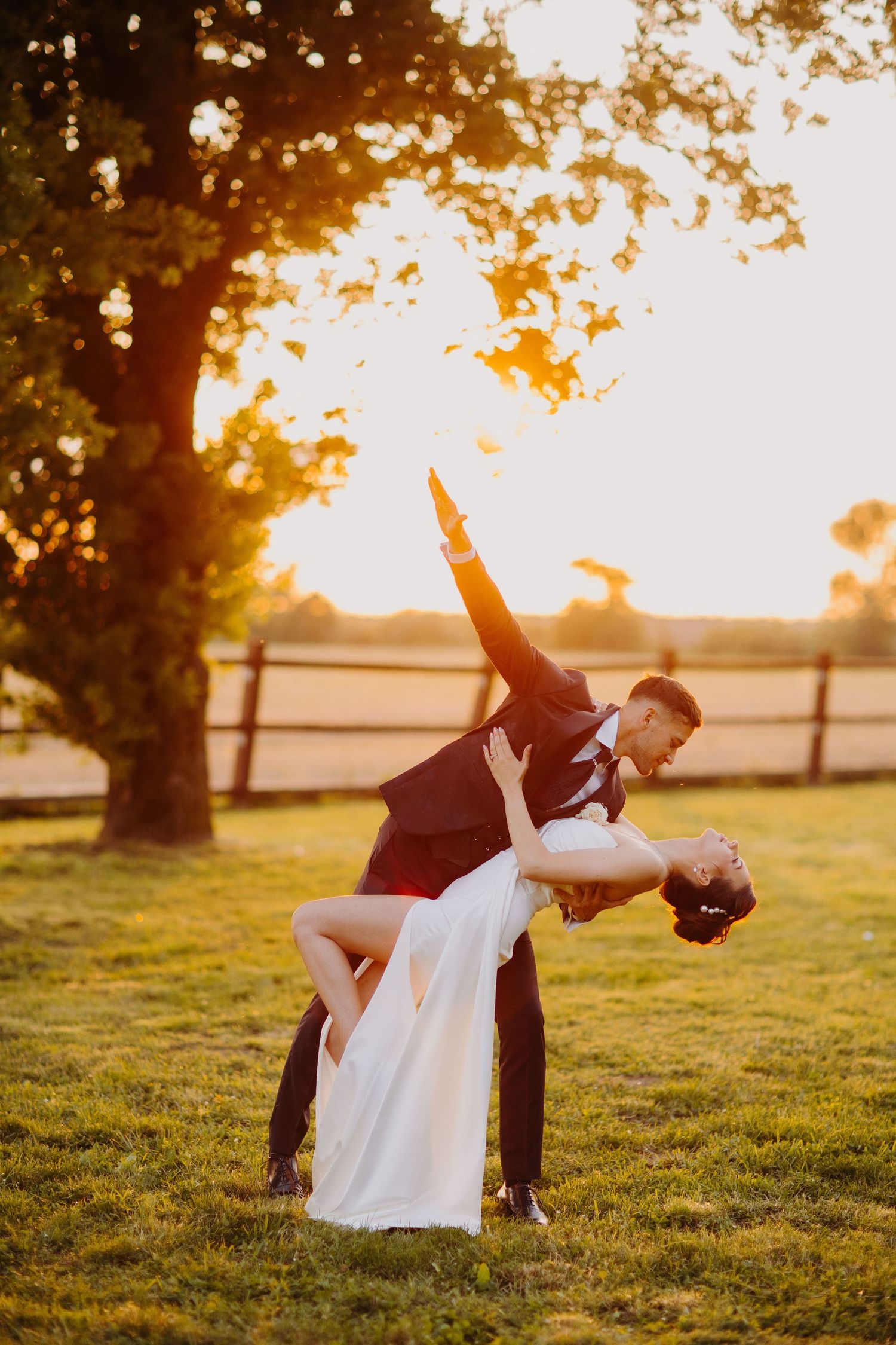 A romantic sunset dip pose captured in a grassy field with golden light filtering through trees in the background.