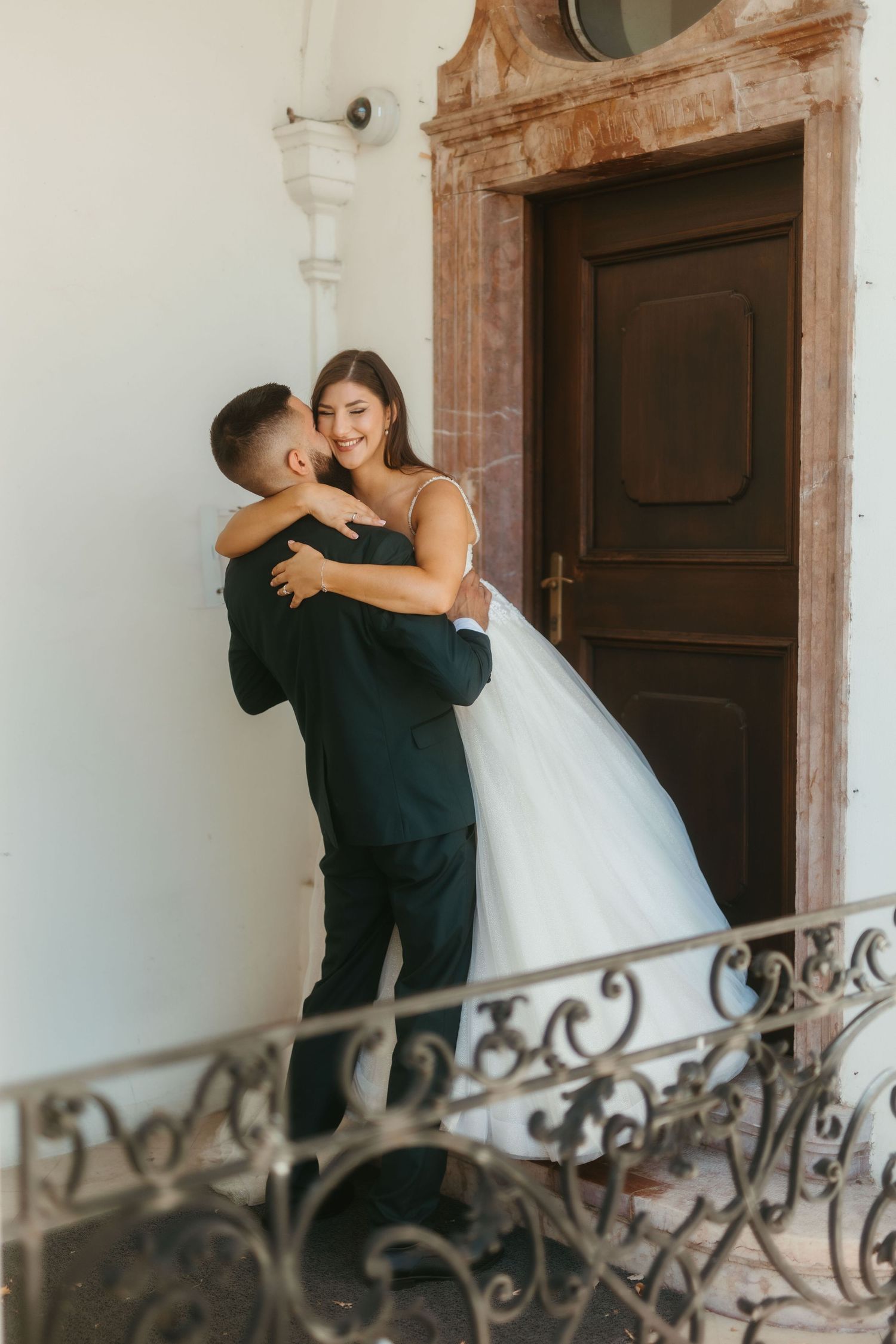 A bride and groom share a romantic moment on an elegant staircase with ornate ironwork railing.