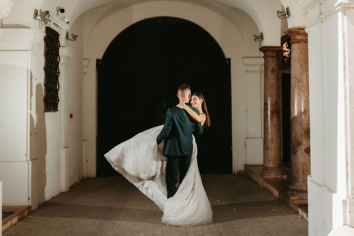 A couple in wedding attire embrace in a romantic kiss while standing in an elegant arched hallway.