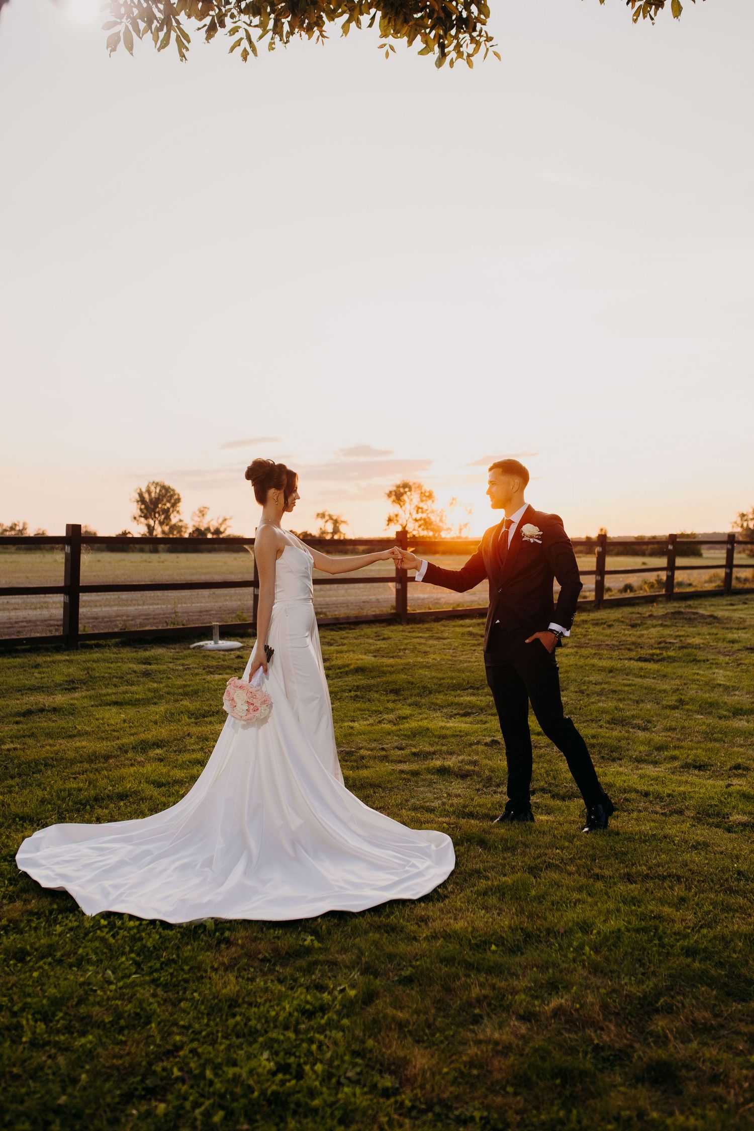 Couple dancing at sunset in a field with a flowing wedding dress creating a romantic silhouette against the golden sky.