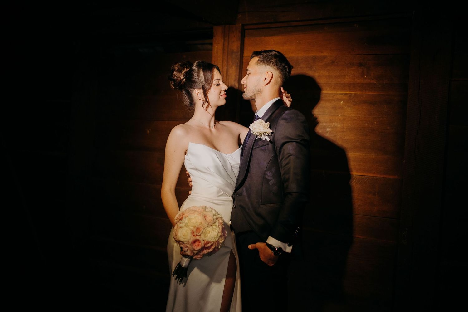 A romantic wedding portrait shows couple embracing in dark wooden interior with dramatic lighting.