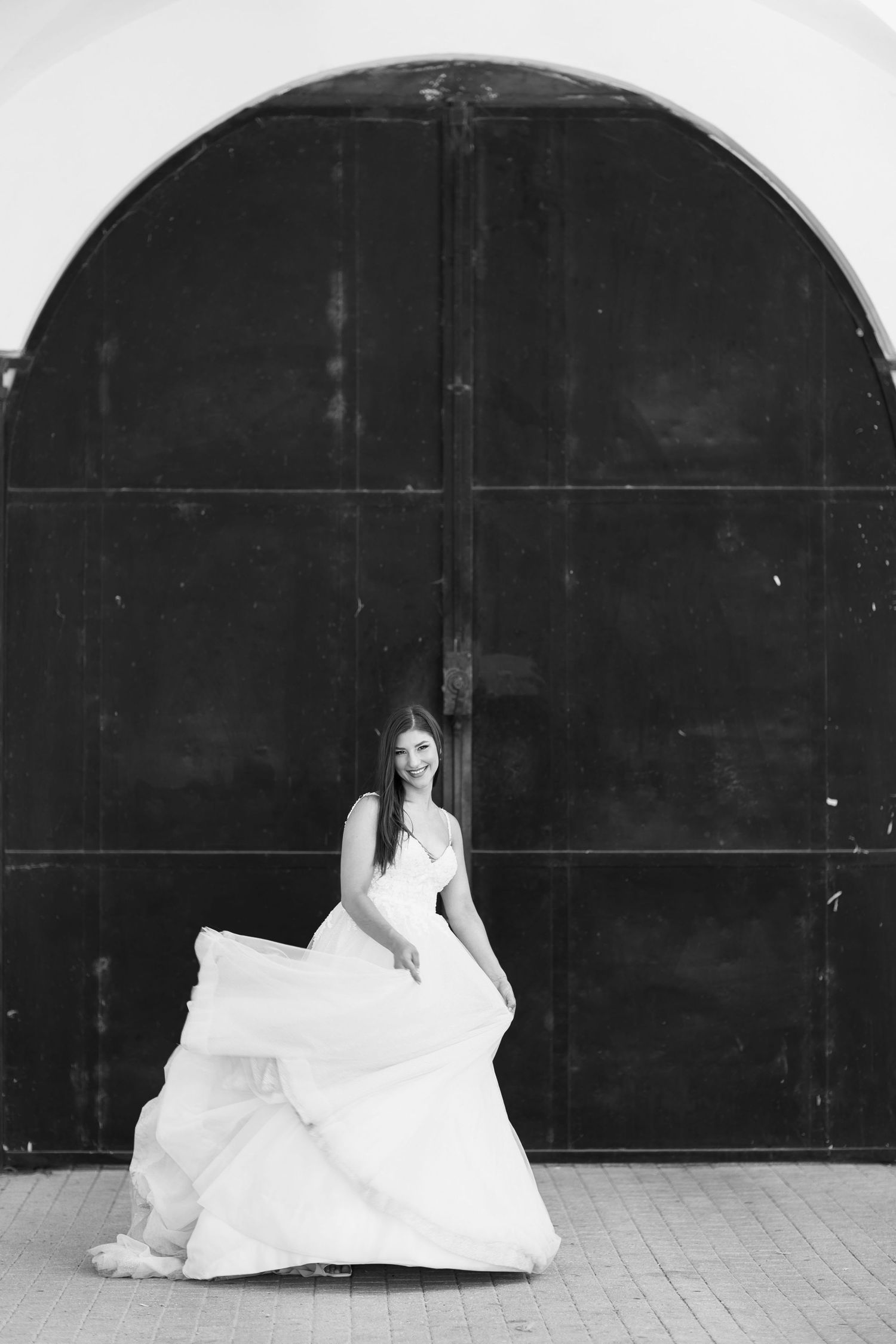 A bride in a flowing white wedding gown poses against a dark arched doorway in a black and white photograph.