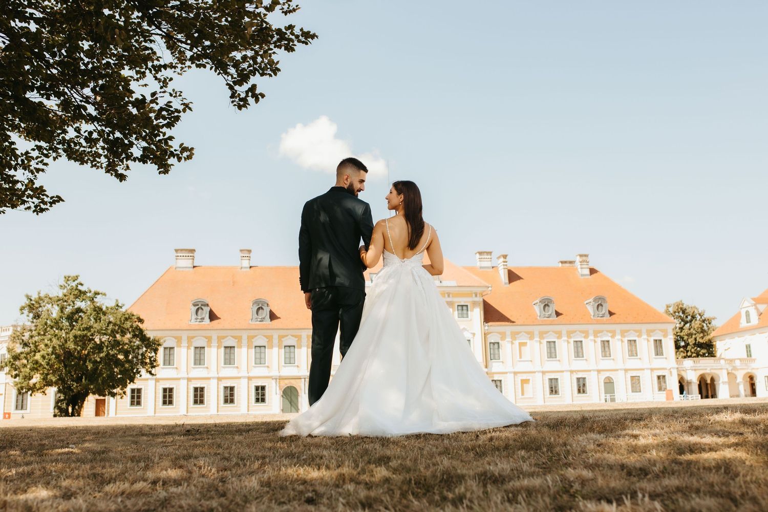 A couple in wedding attire poses in front of a grand mansion with orange roof and white walls on sunny day.