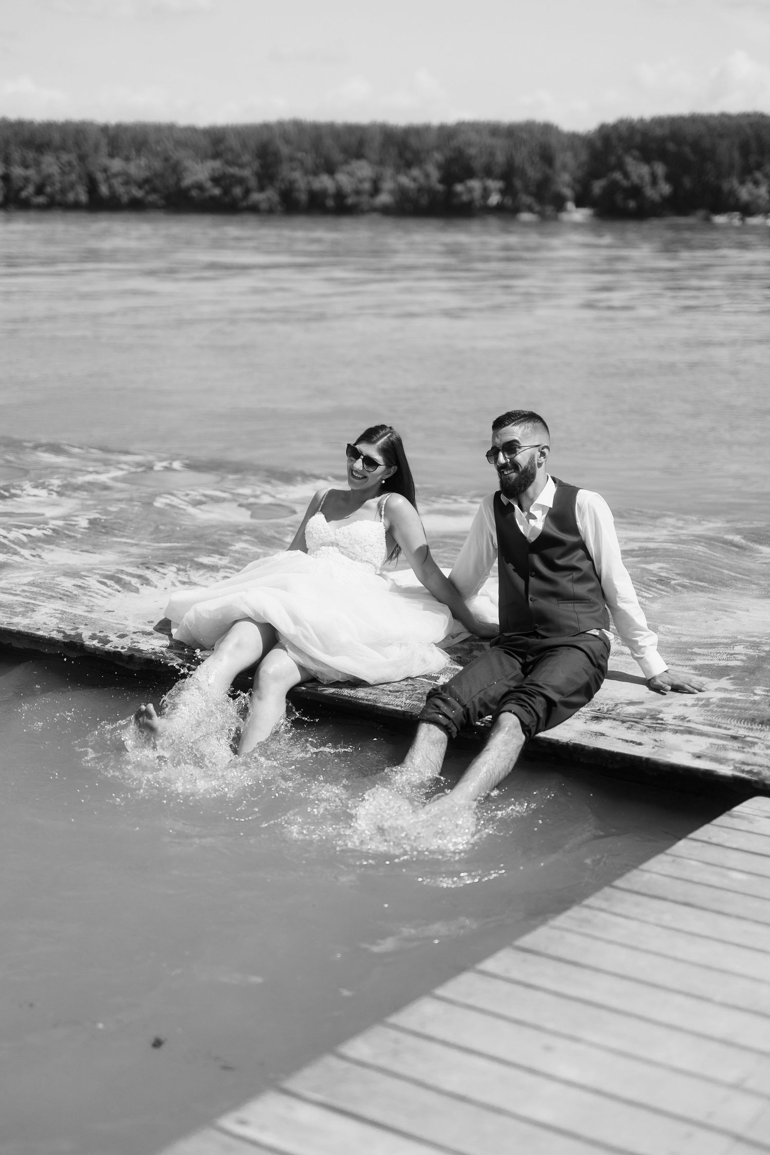 A couple in wedding attire sitting on a dock with their feet in the water, captured in black and white.