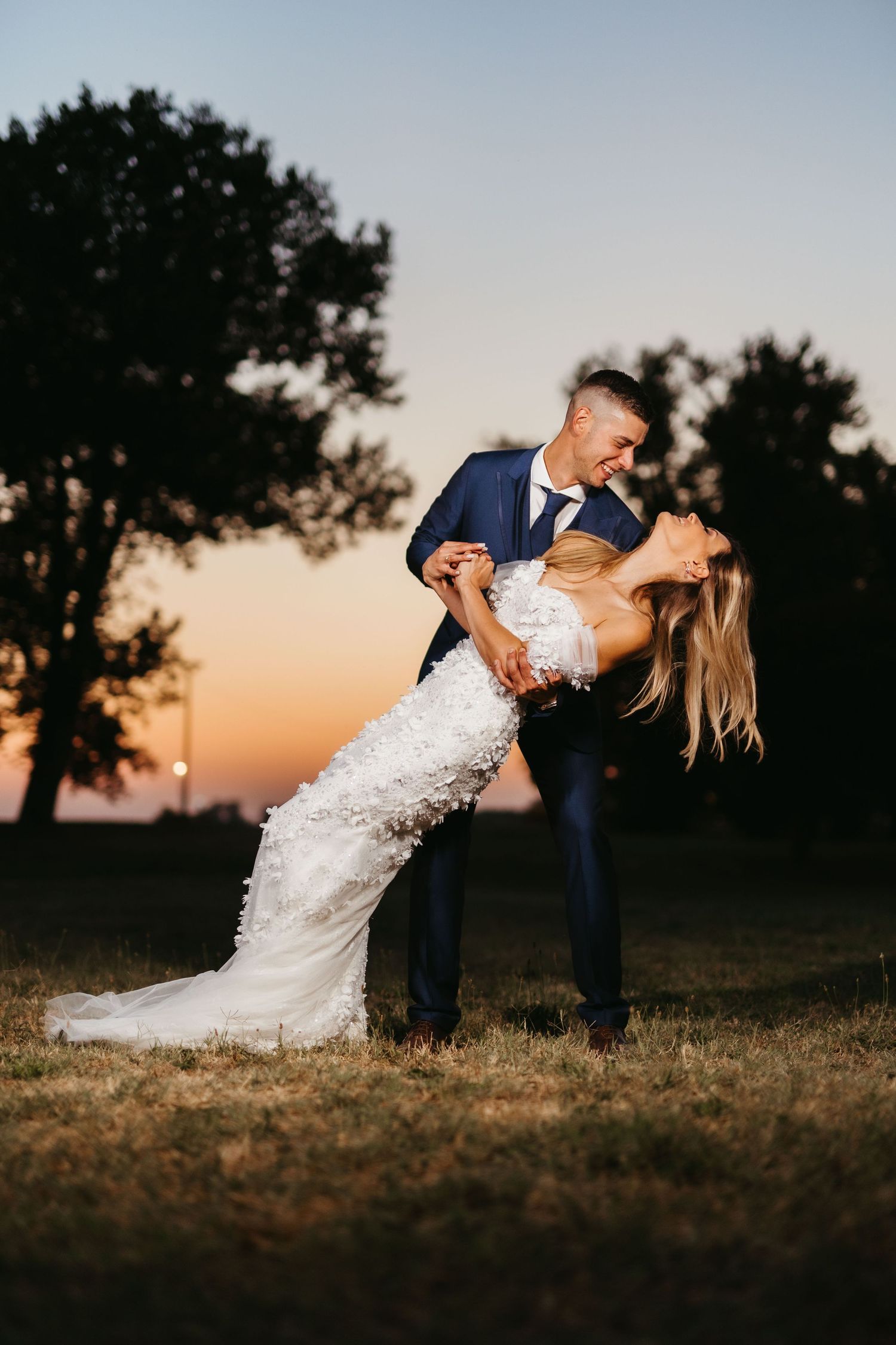 Dramatic dip pose wedding photo of couple against sunset sky in formal wedding attire.