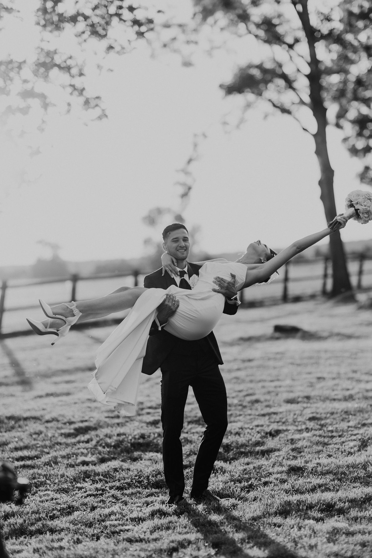 Black and white photo of a newlywed couple in joyful embrace on grassy field during outdoor wedding celebration.