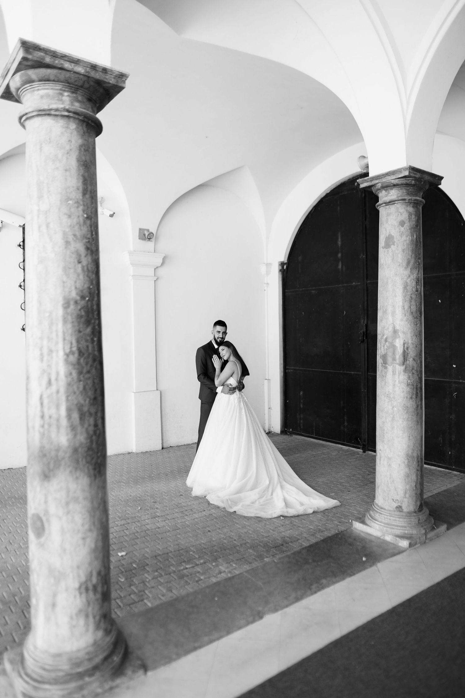 A romantic black and white wedding photo captured between elegant stone columns in an arched hallway.