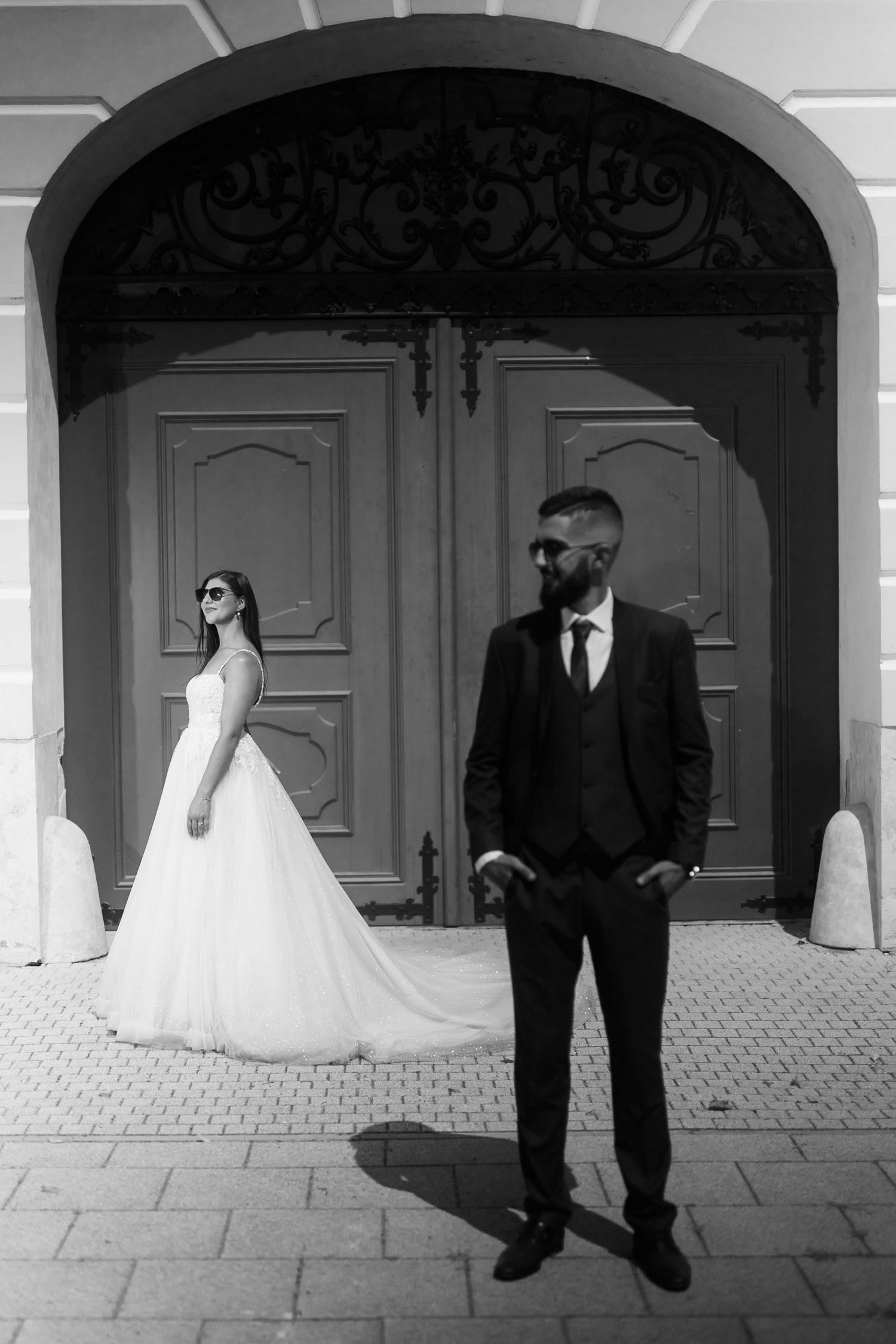 Black and white wedding photo with bride in flowing white gown and groom in dark suit standing by ornate doorway entrance.
