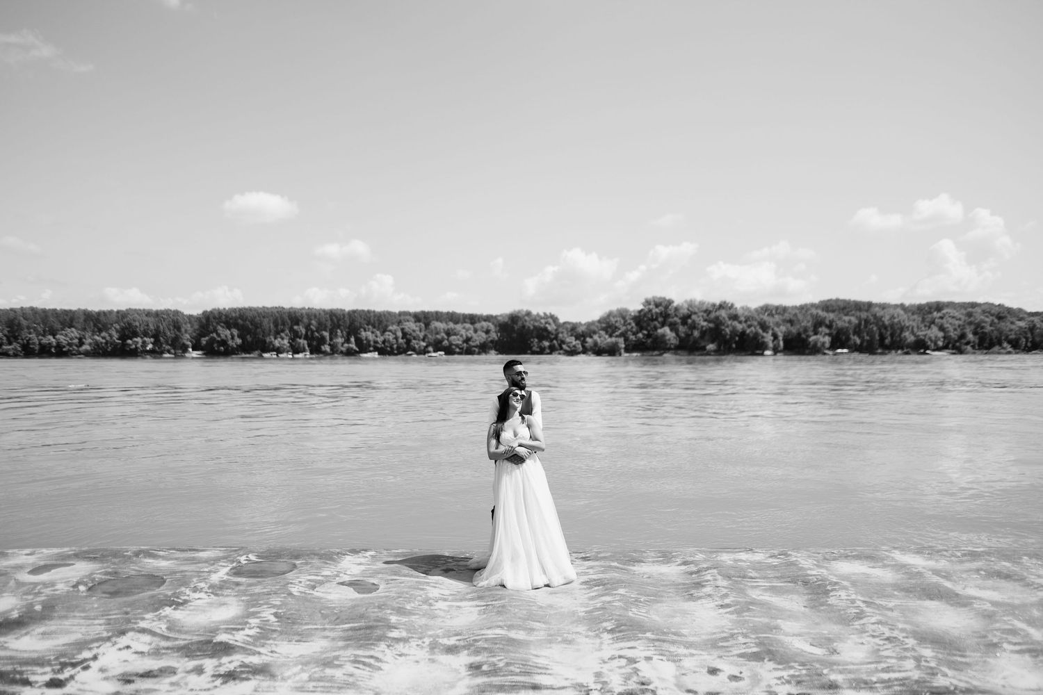 A lone figure in a white dress stands at the edge of a lake with trees lining the distant shoreline in black and white.