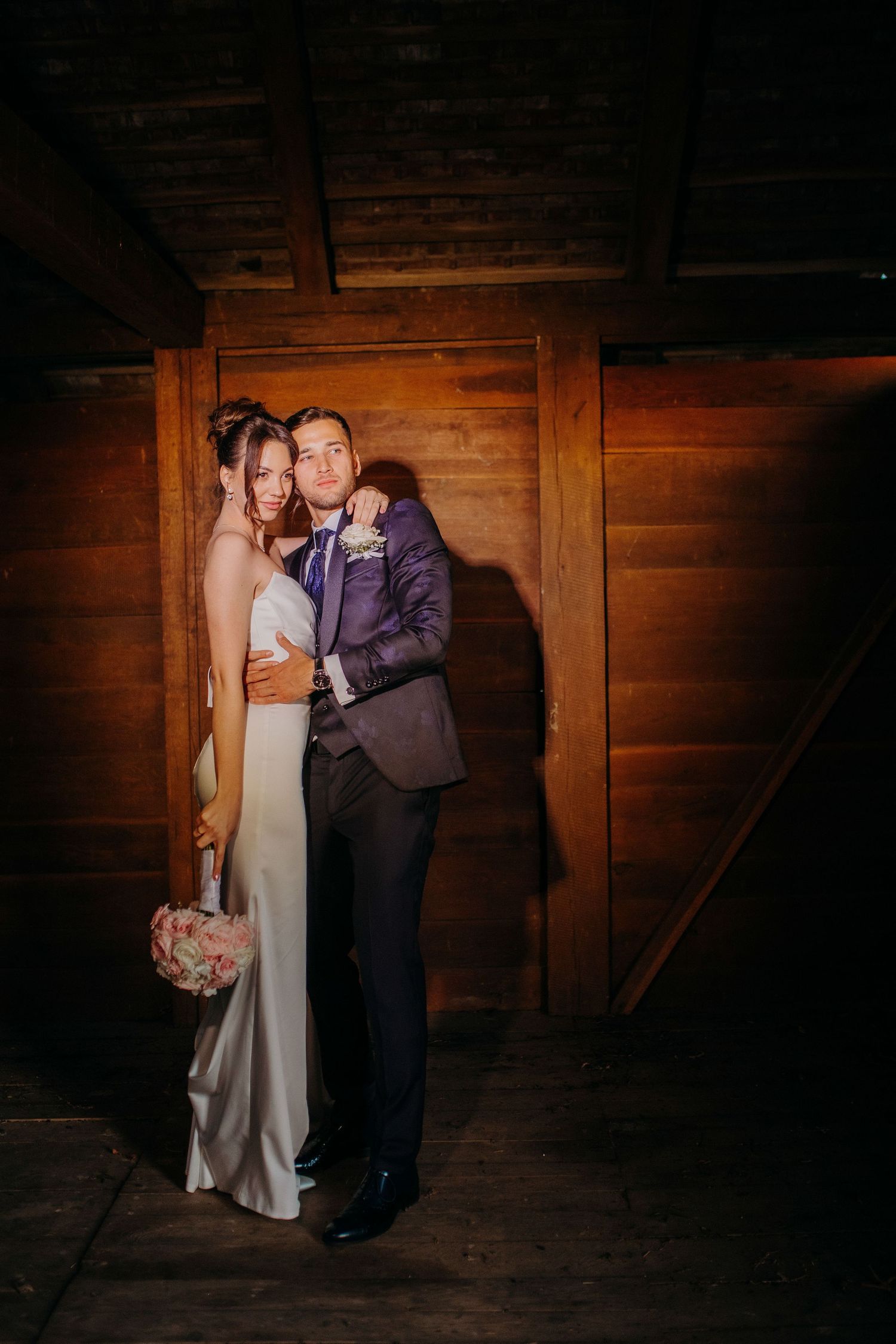 Romantic wedding couple poses together in rustic wooden barn setting with dramatic warm lighting.