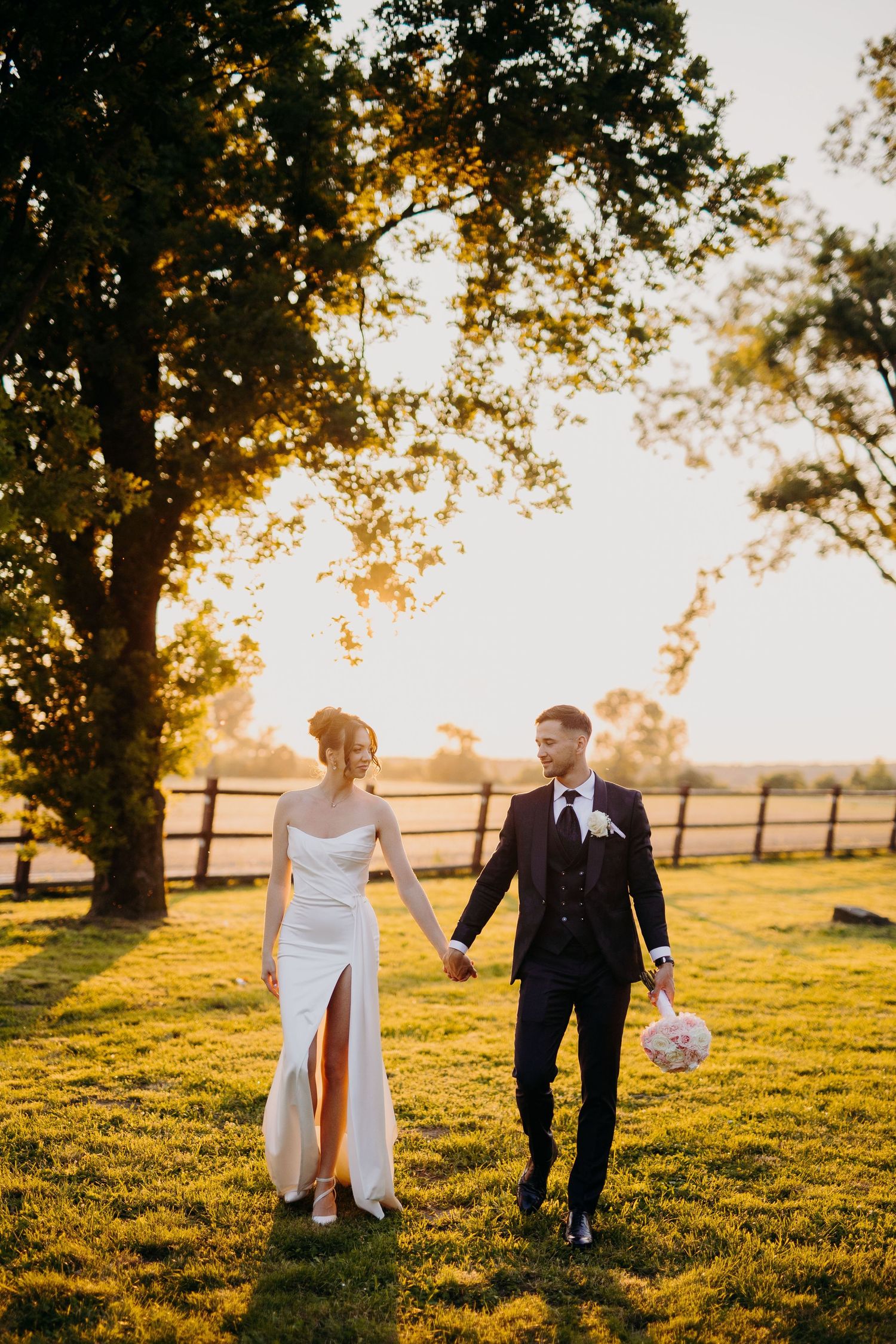 A couple walks hand in hand through a sunlit field at golden hour, with wooden fences and trees in the background.