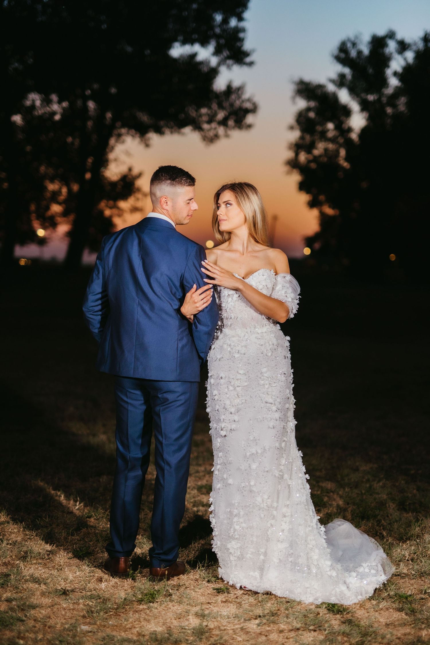 A couple shares an intimate moment at sunset, with the bride in white lace and groom in navy suit.