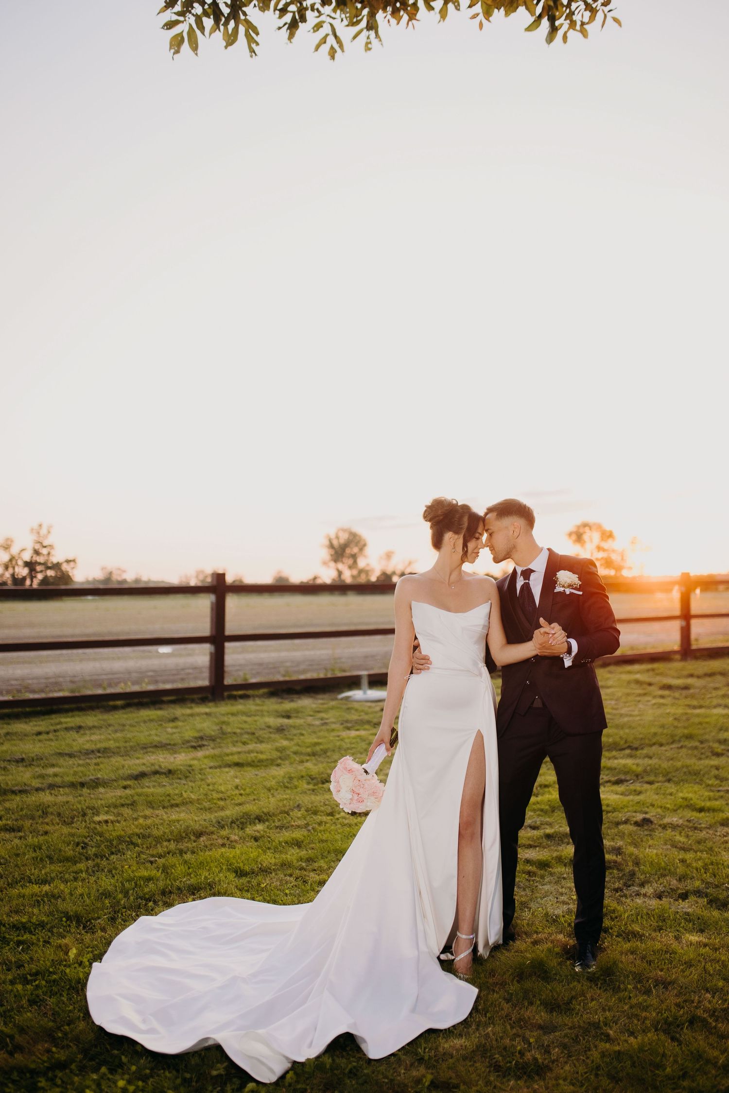 A romantic sunset wedding photo of a couple in formal attire standing in a grassy field with a wooden fence.