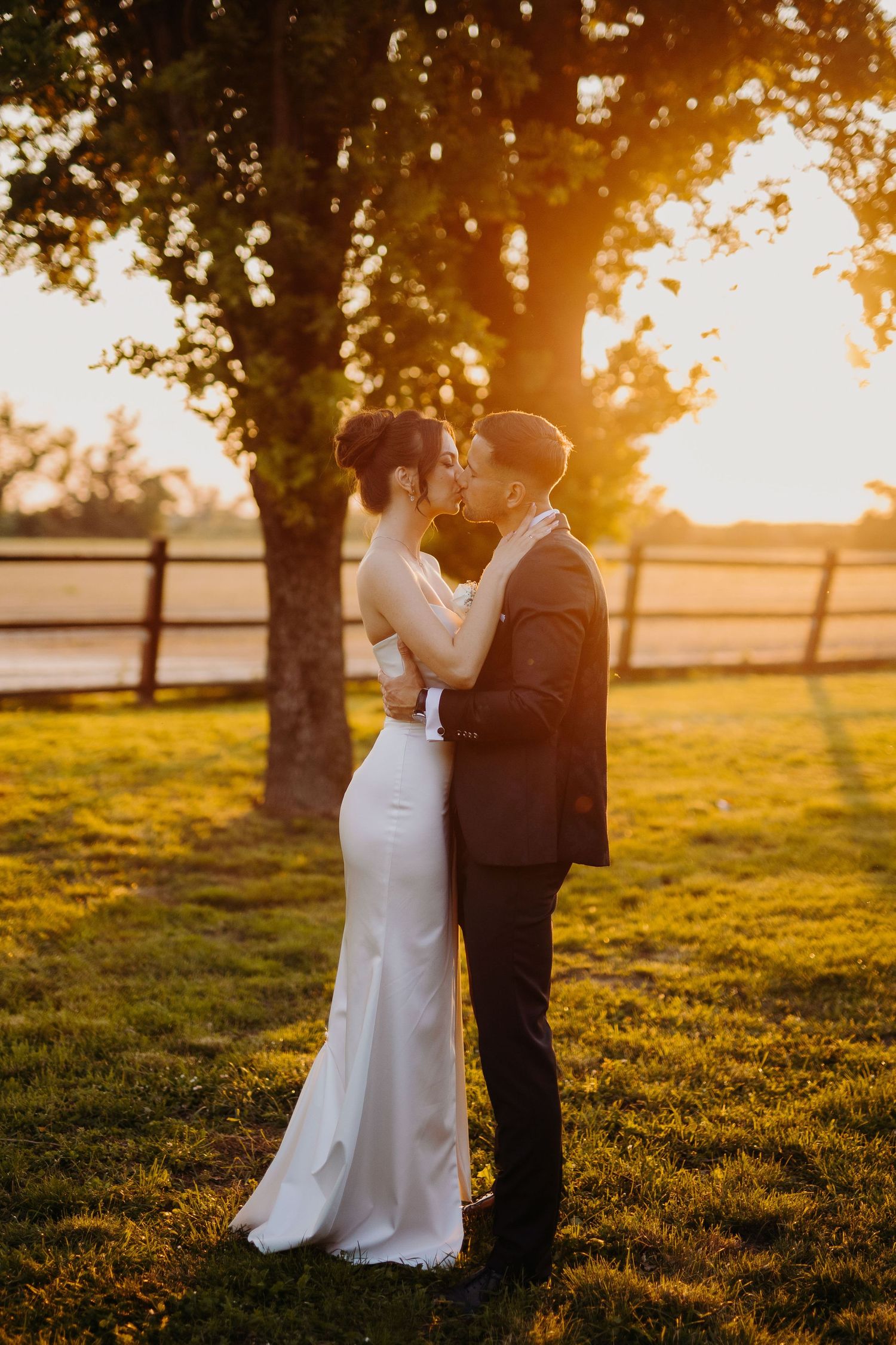 A romantic sunset wedding photo of a couple embracing outdoors with golden light filtering through trees.