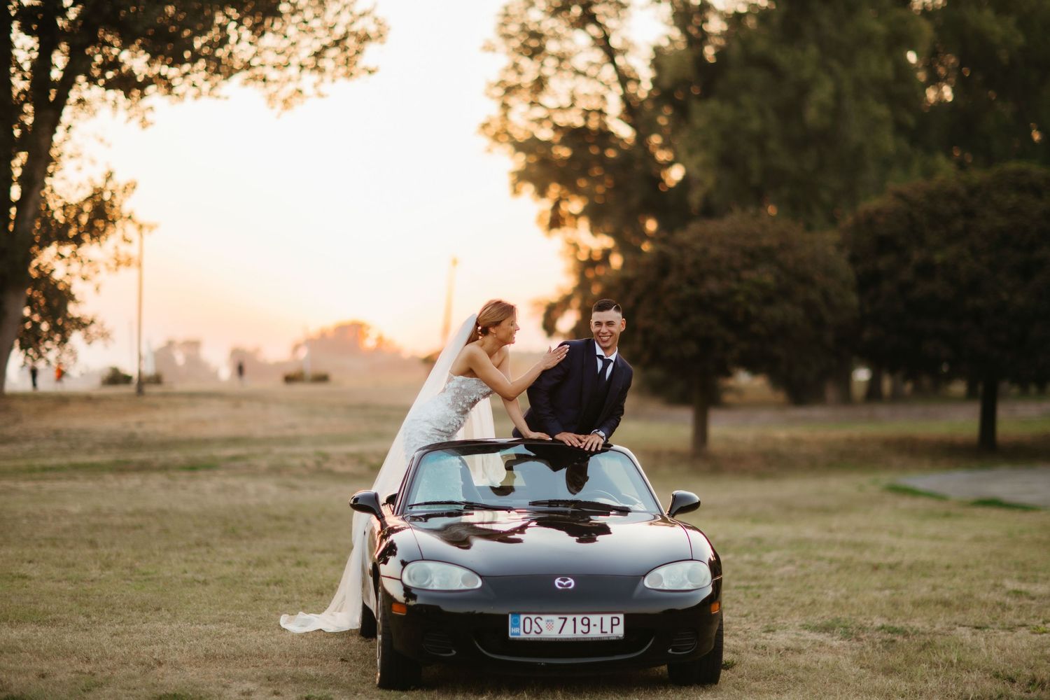 Wedding couple stands through convertible roof of black sports car during sunset photoshoot.