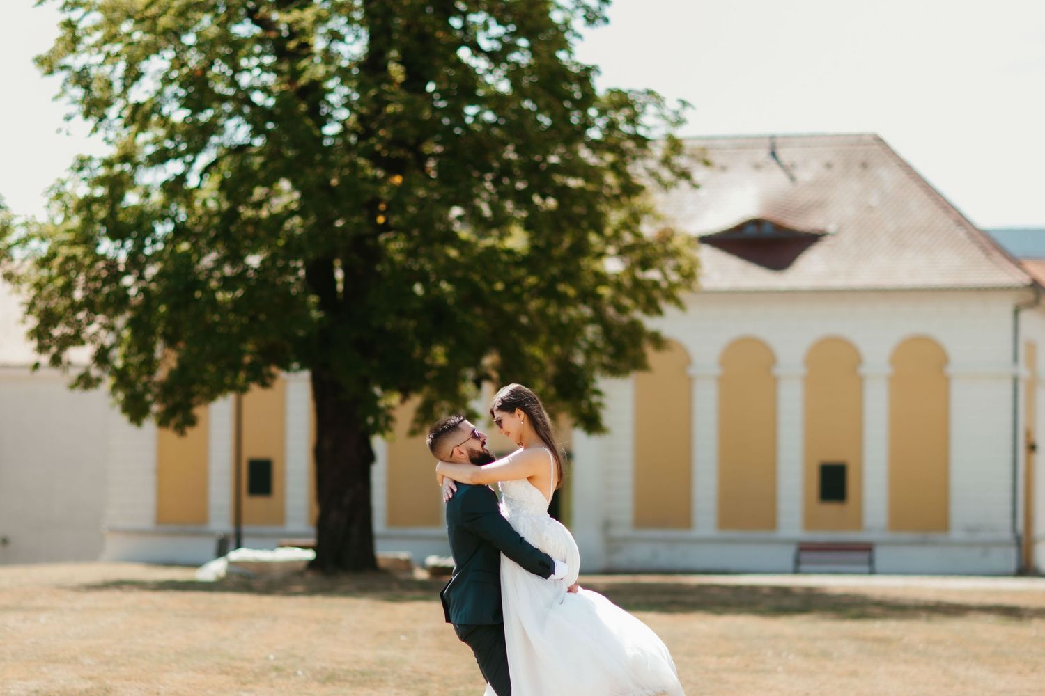 Romantic embrace between newlyweds in front of a yellow church building with a large tree on a sunny day.