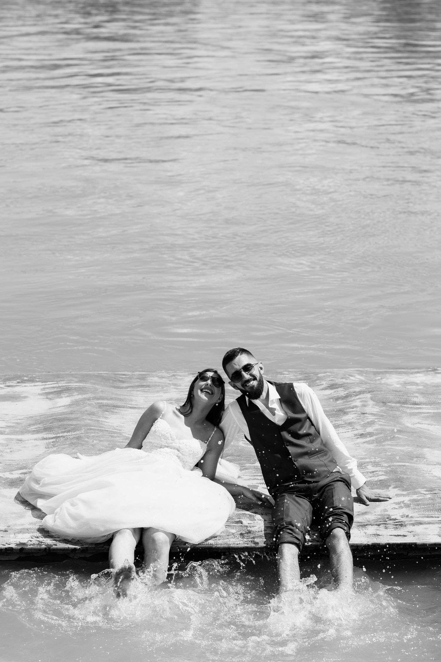 A joyful couple in wedding attire sits at the edge of water, splashing their feet and smiling together in black and white.