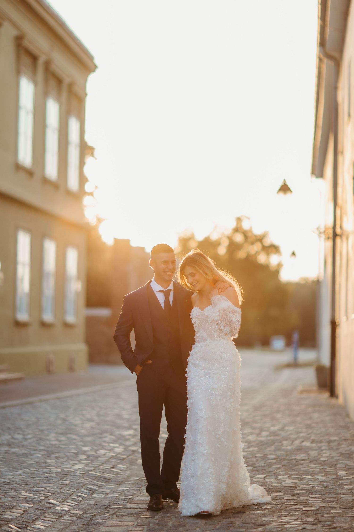 Sunlit wedding portrait with couple on cobblestone street between historic buildings.