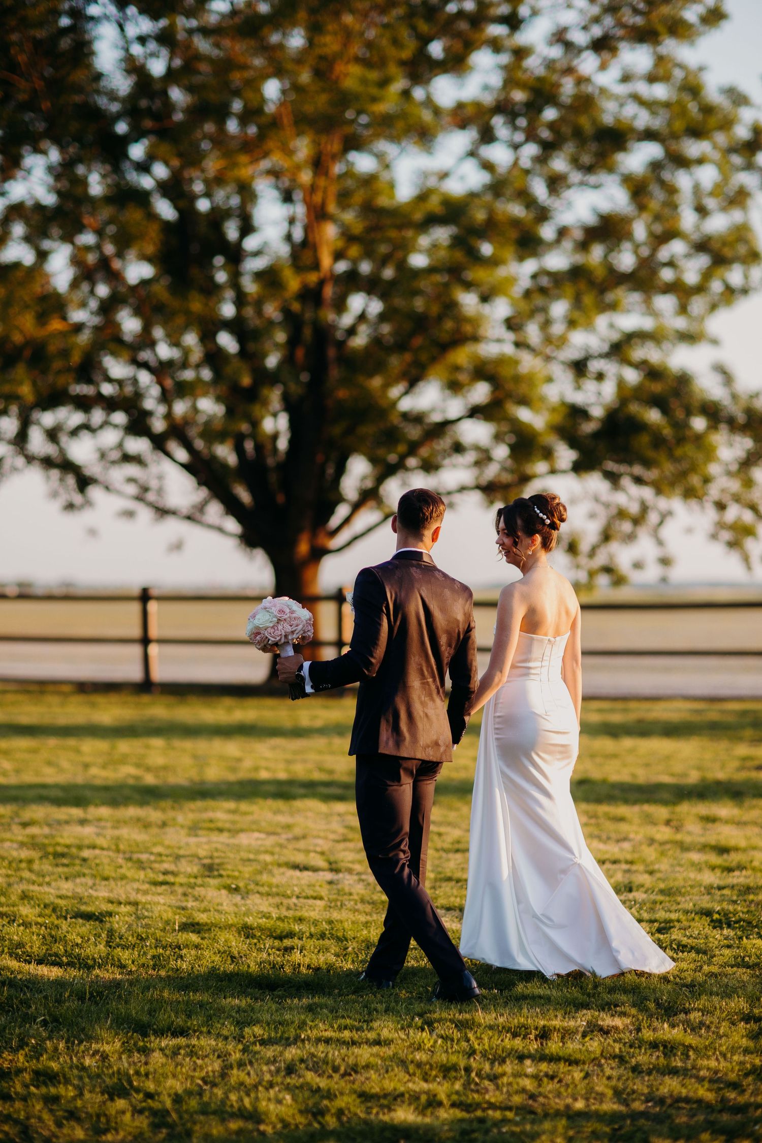 A couple walks hand in hand across a sunlit grassy field during golden hour wedding photography session.