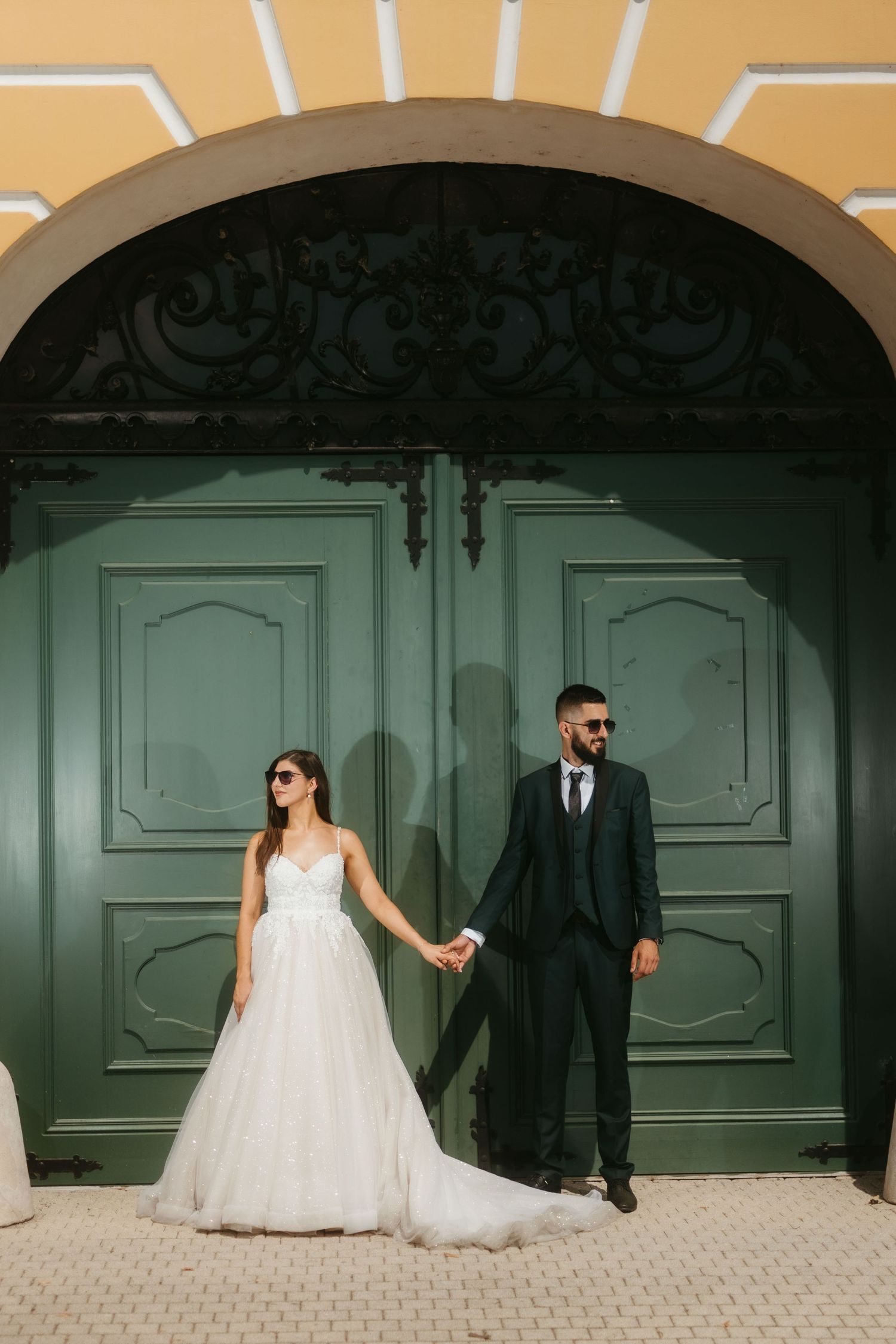A couple in wedding attire poses against large mint green doors with a decorative arched entrance.