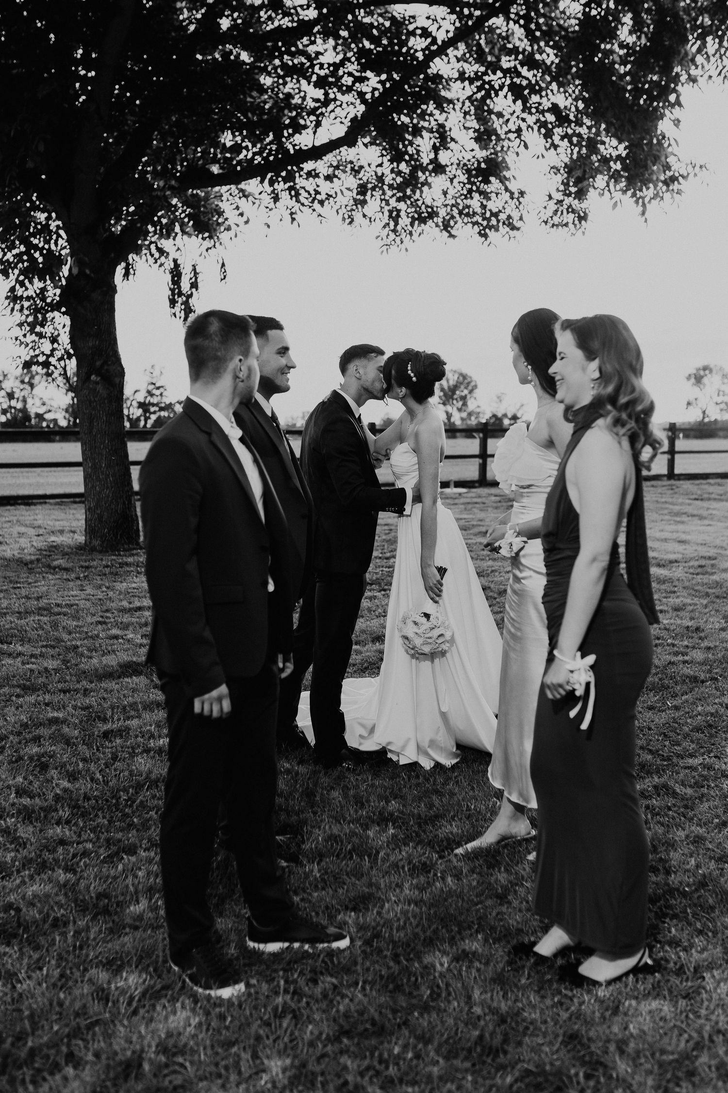 Black and white wedding party candid photo sequence showing group gathering under a tree in an outdoor setting.