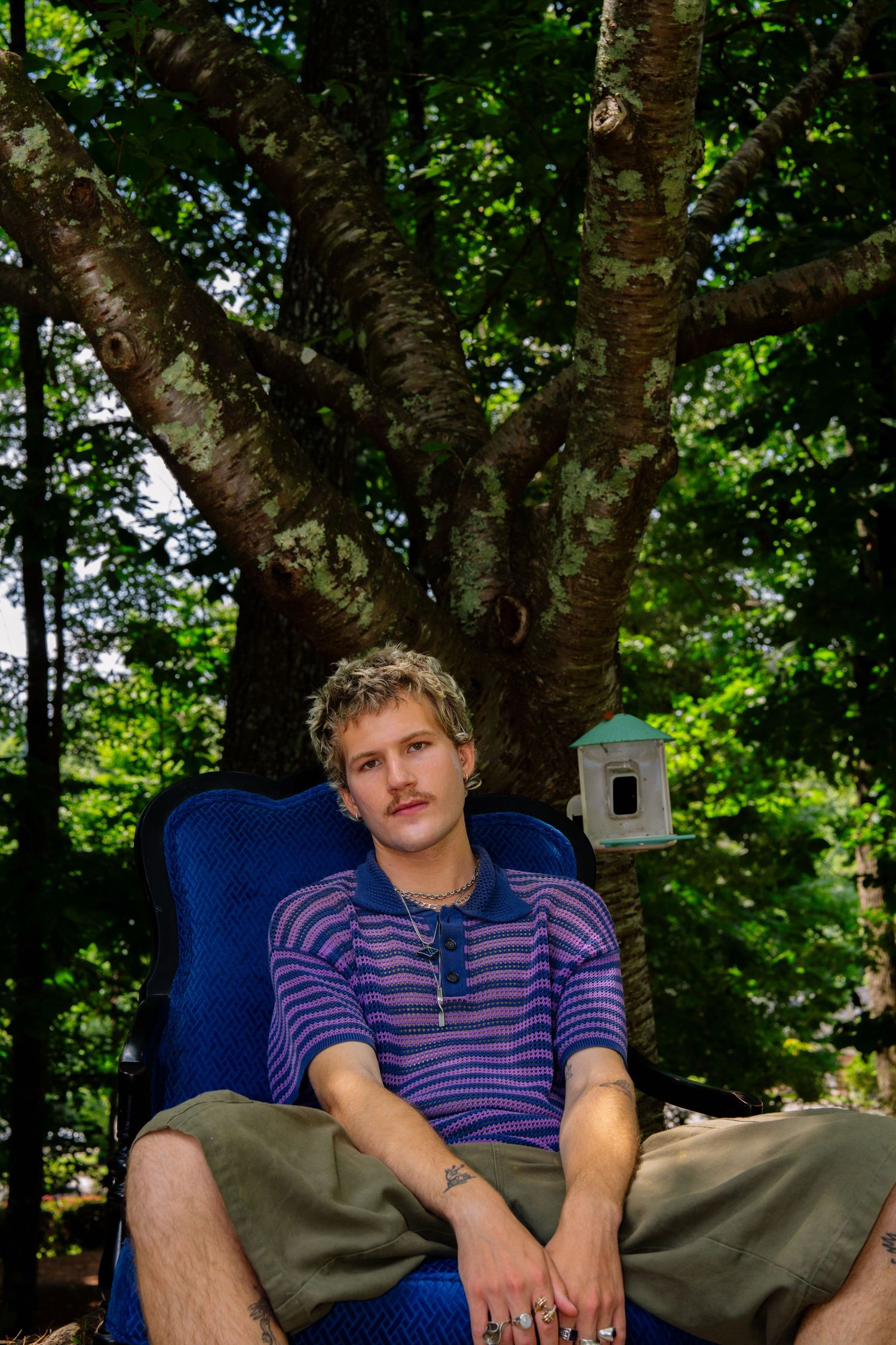 Casual outdoor portrait with person lounging in blue chair against green forest backdrop.