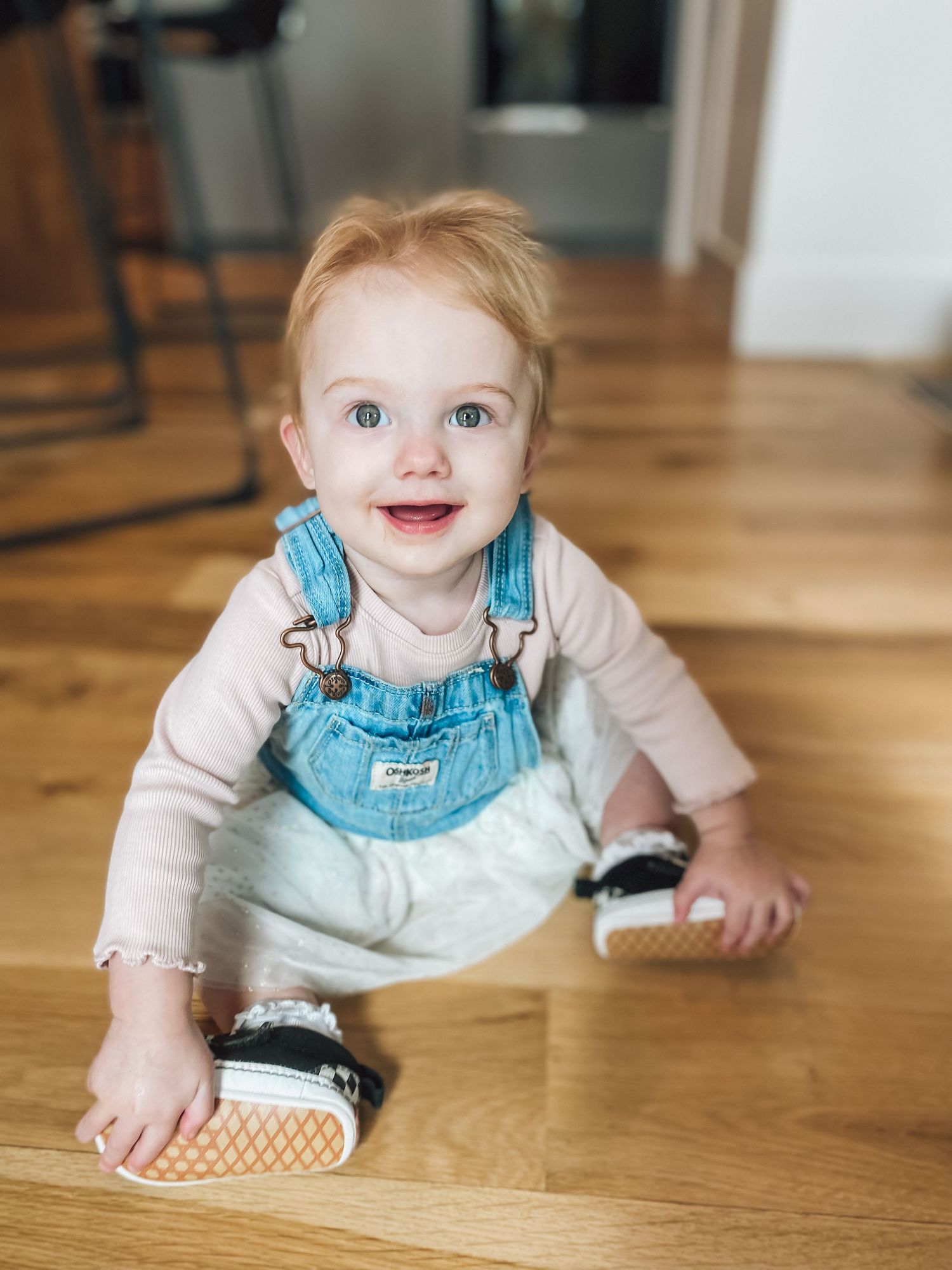 Baby in denim overalls sits on hardwood floor with checkerboard shoes.