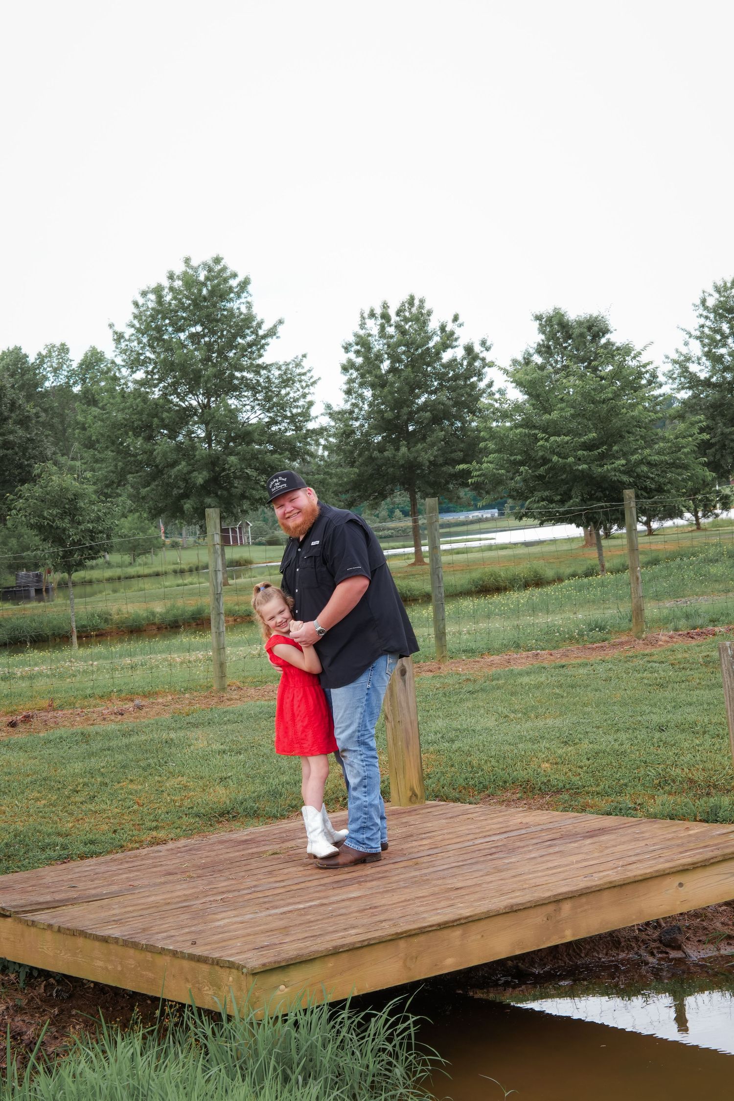 Two people sharing a tender moment on a wooden dock by a pond in a pastoral setting.