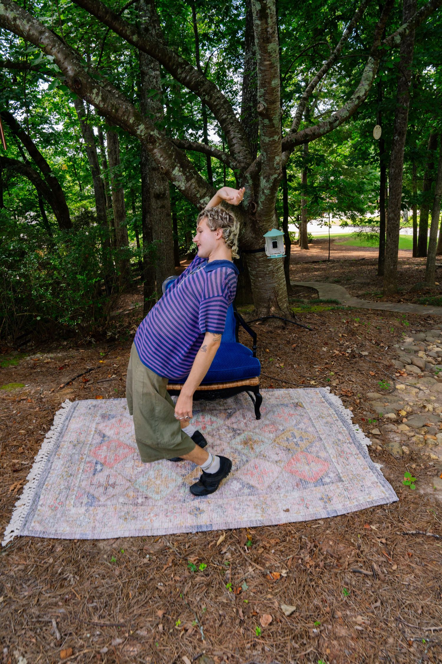 Person in striped shirt kneels on patterned rug under trees outdoors.