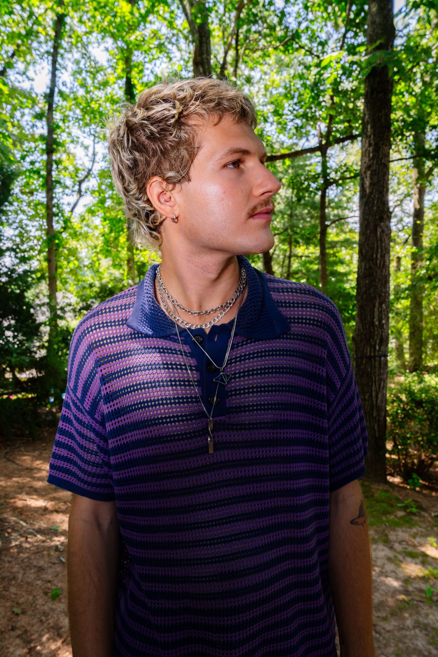 Person wearing a purple and pink striped polo shirt poses in a wooded outdoor setting.
