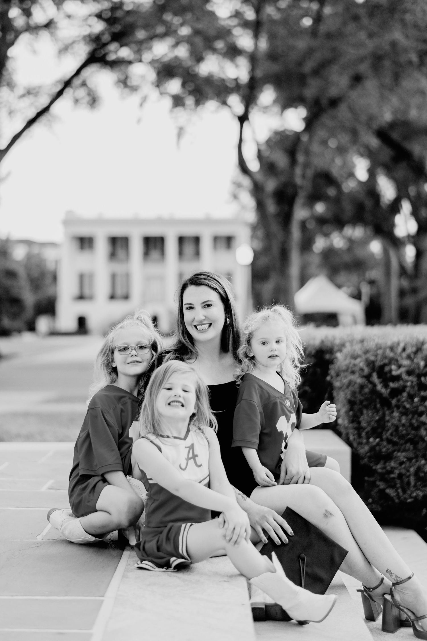 Black and white portrait of a family sitting together and smiling on outdoor steps near a building with trees in background.